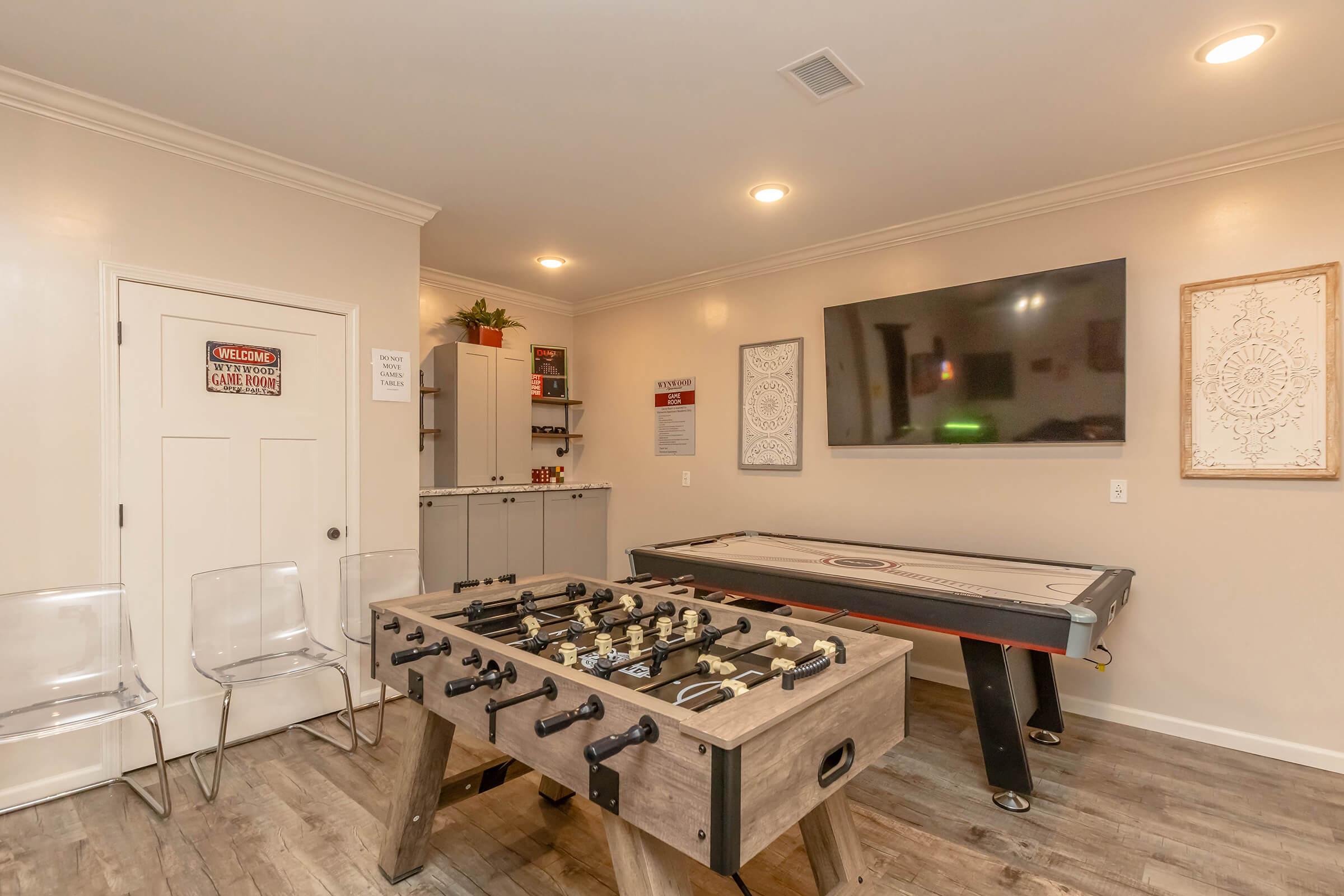 A cozy game room featuring a foosball table and an air hockey table, with a flat-screen TV mounted on the wall. There are decorative wall art pieces, a small cabinet, and transparent chairs. Soft lighting adds to the inviting atmosphere of the space.