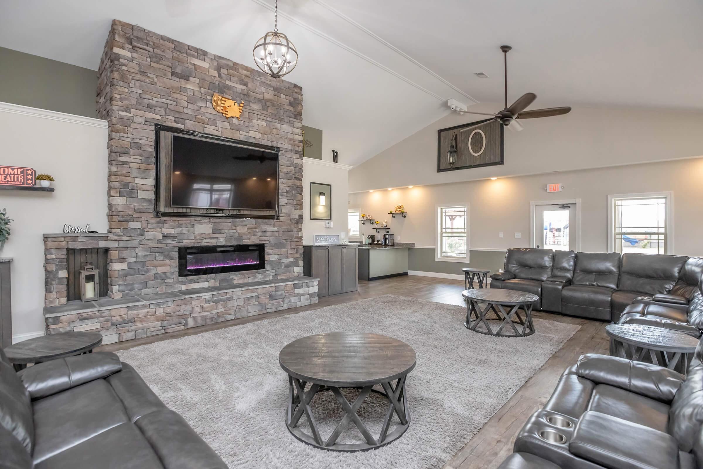 A stylish living space featuring a stone fireplace with a mounted TV, modern furniture including dark leather sofas, a round coffee table, and large windows. The decor includes a contemporary chandelier and soft lighting, creating a cozy and inviting atmosphere.
