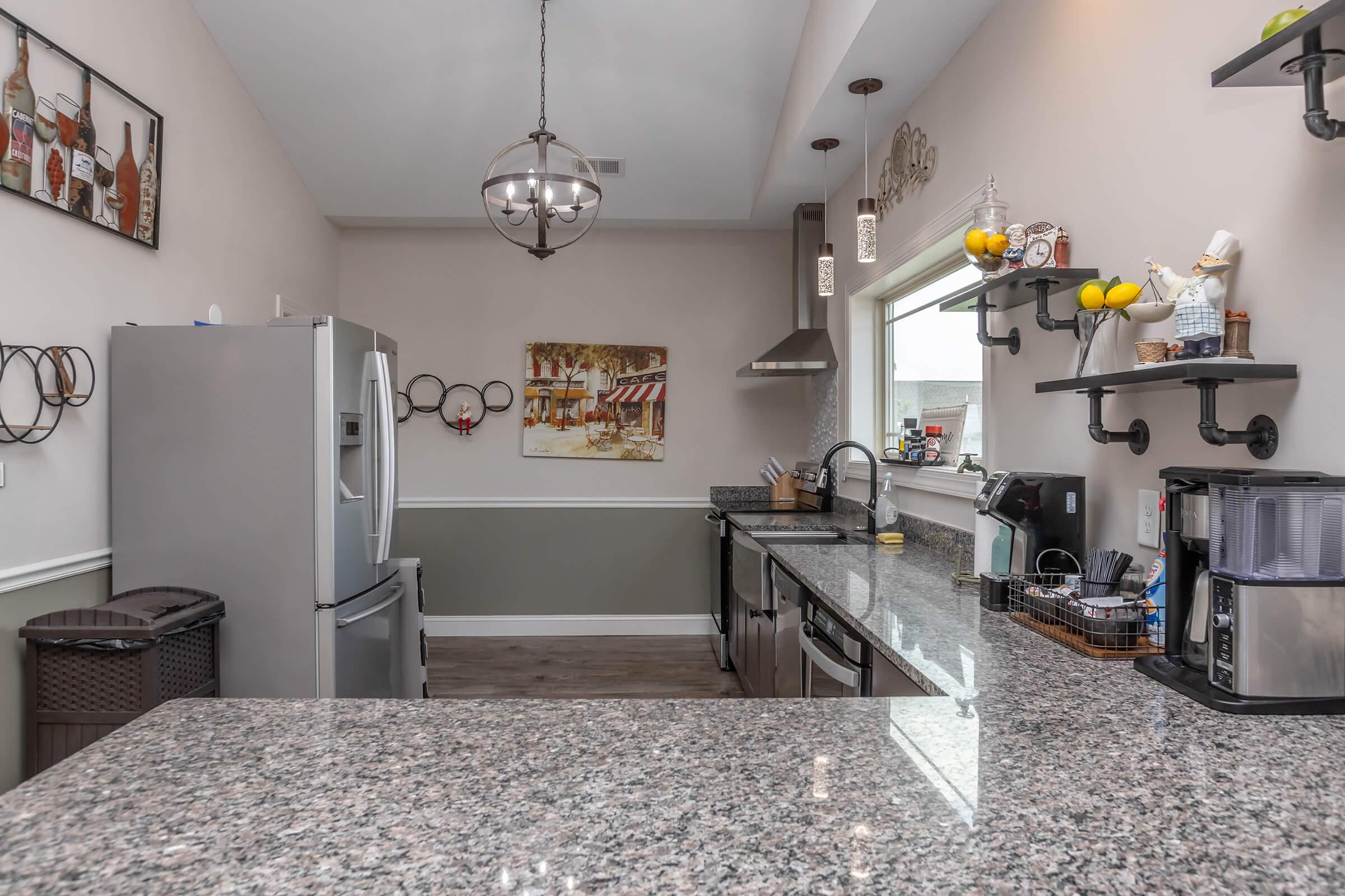 Modern kitchen featuring stainless steel appliances, including a refrigerator and stove, with granite countertops. Decorated with wall art, a fruit bowl, and open shelves displaying kitchen items. The area is well-lit with pendant lighting and includes a neutral color scheme.