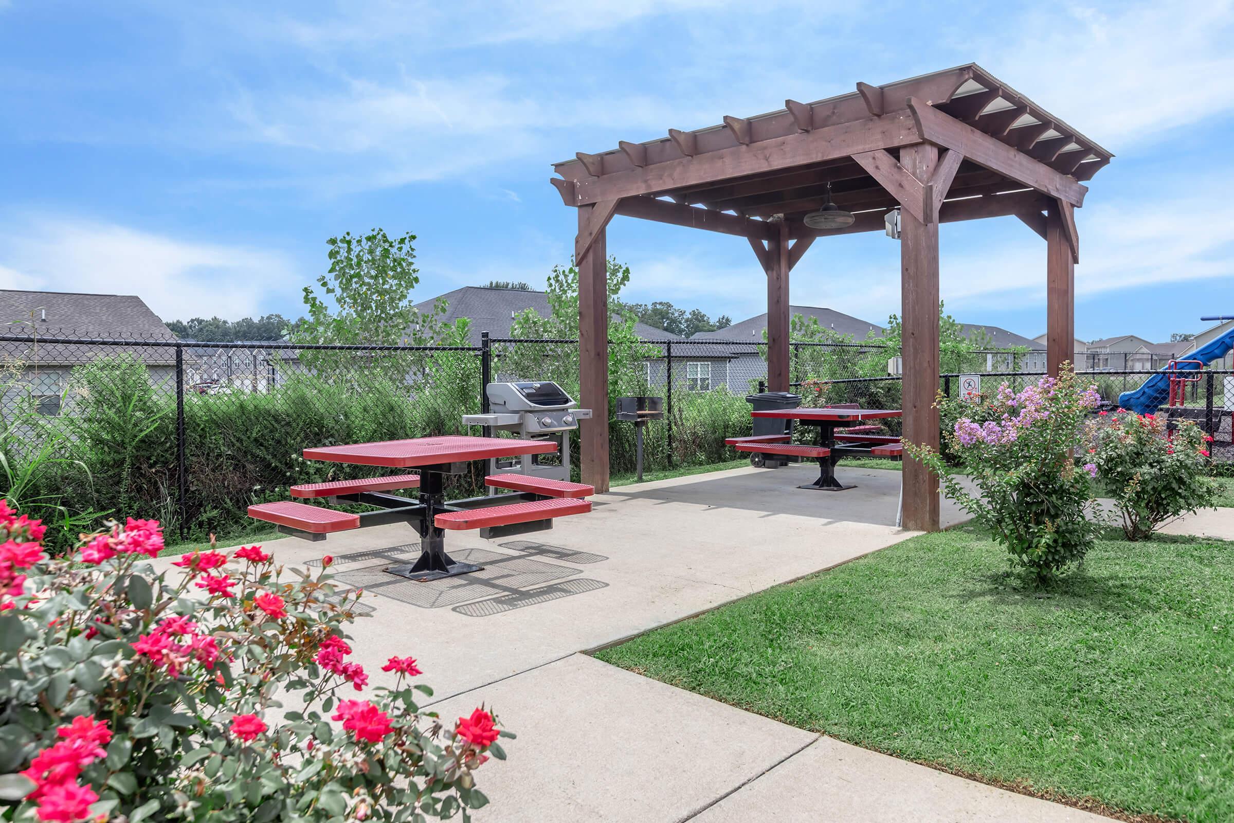 A shaded outdoor picnic area featuring red tables under a wooden pergola. There is a grill nearby and landscaped gardens with blooming pink roses. In the background, there are residential buildings and a playground visible beyond the fence. The sky is clear and blue.