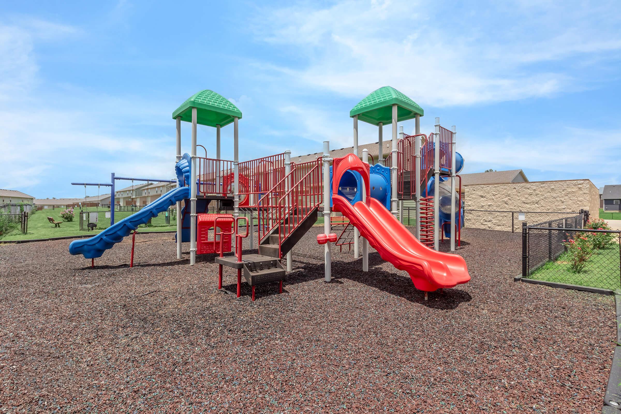 A colorful playground with two slides, climbing structures, and a connected play area. The playground is surrounded by a fence, with grass and a few buildings visible in the background under a clear blue sky.