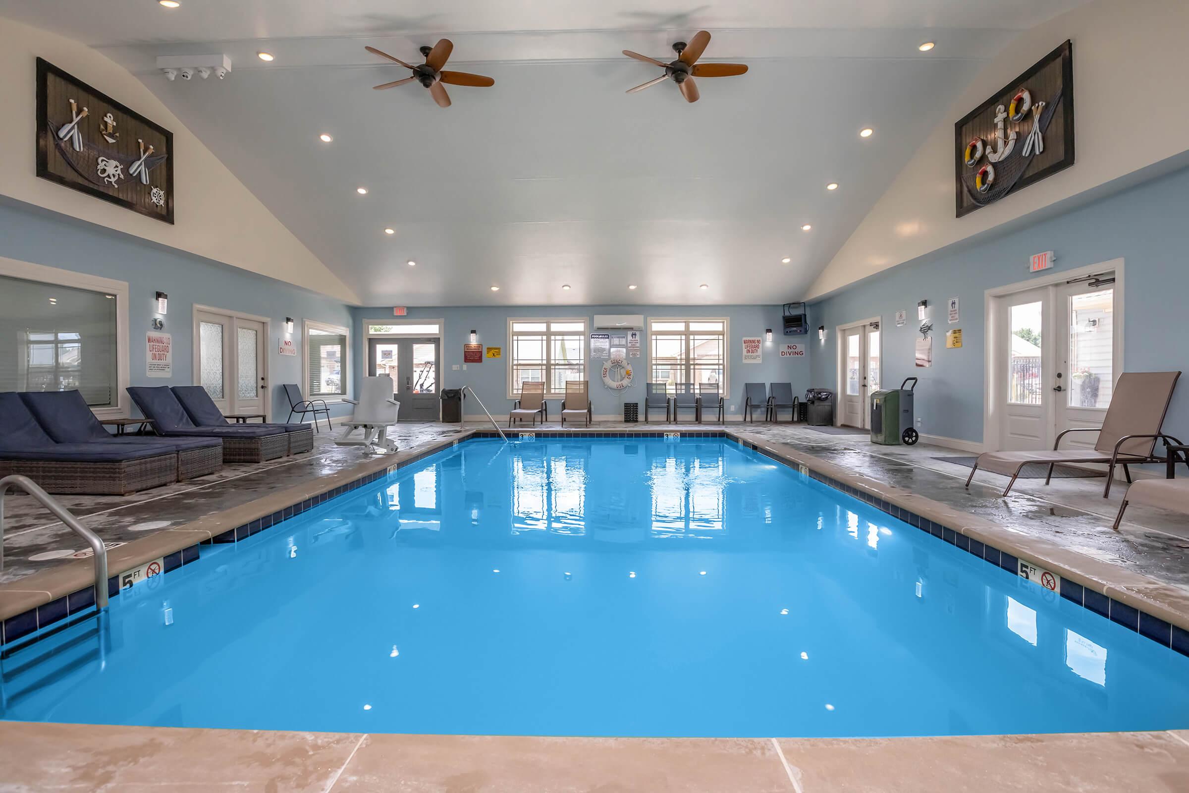 Indoor swimming pool with clear blue water, surrounded by lounge chairs. Ceiling fans are installed above, and large windows provide natural light. The walls are painted light blue, with decorative signs and safety notices displayed. The area appears clean and inviting, perfect for relaxation.