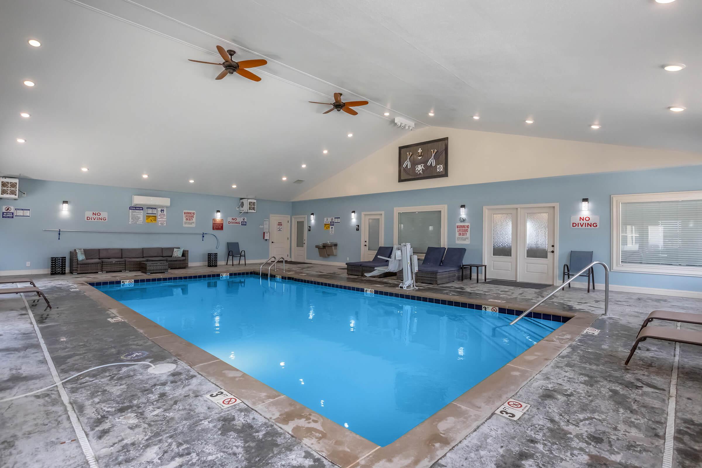 Indoor swimming pool area featuring a well-lit space with ceiling fans and a mix of lounging chairs and poolside seating. The pool is surrounded by safety signage, and there are steps leading into the water. The walls are painted light blue, creating a relaxing atmosphere.