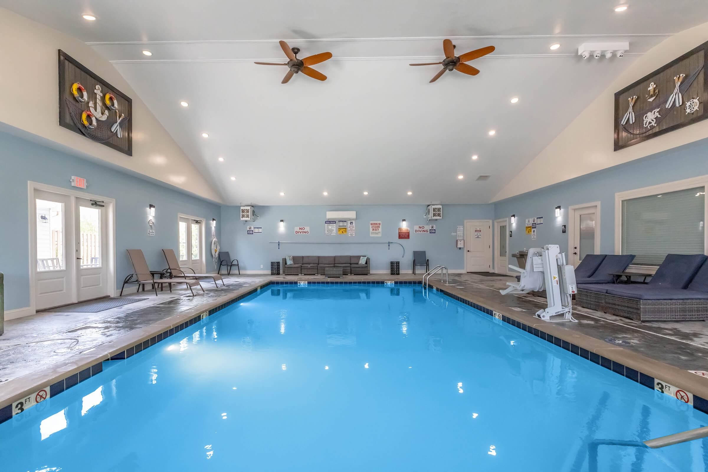 Indoor swimming pool area with bright blue water, surrounded by lounge chairs and decorative wall art. Ceiling fans provide air circulation in the spacious room, which is well-lit with natural light coming from large windows and doors. Safety signs are posted around the pool.