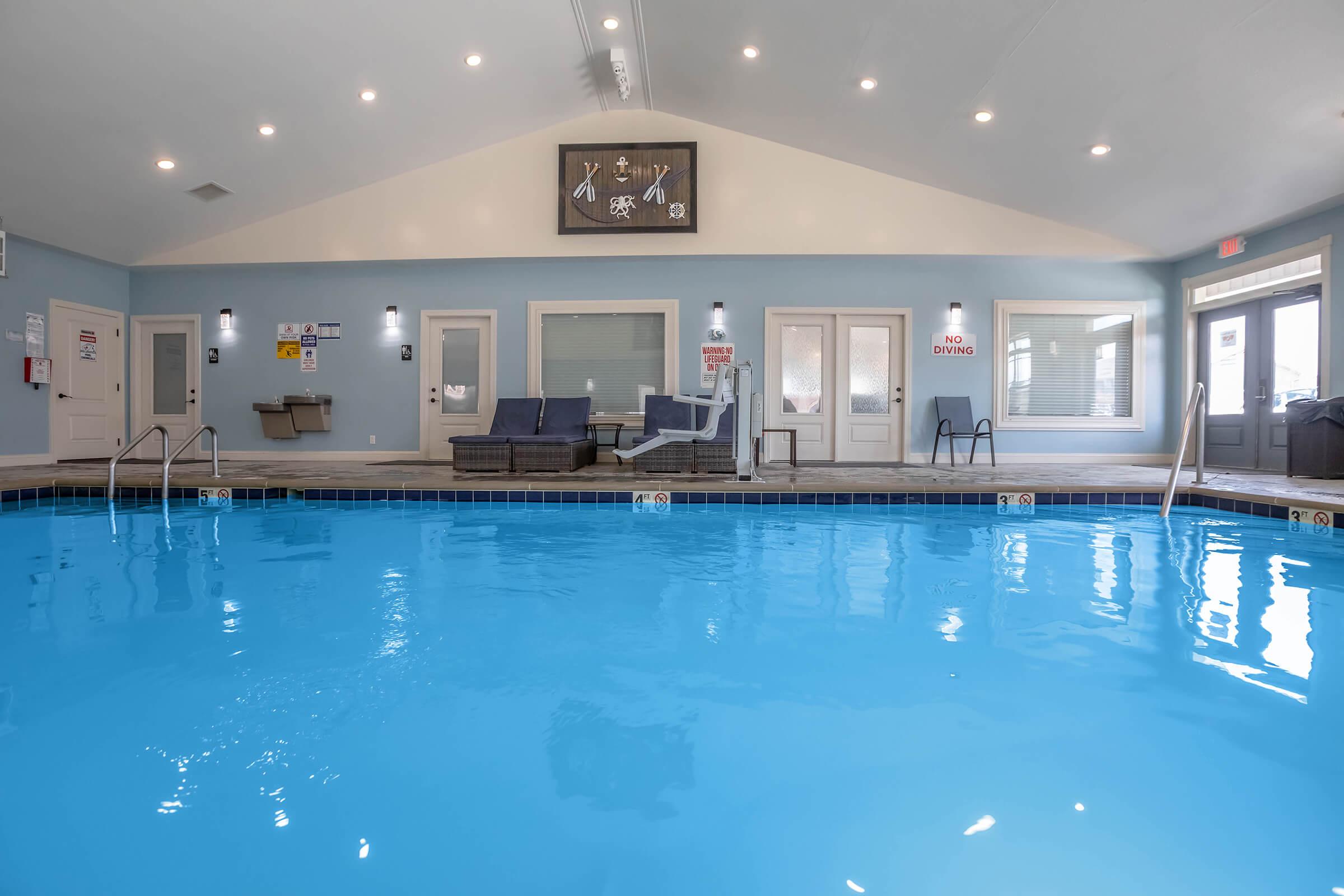 Indoor swimming pool with clear blue water and comfortable lounge chairs nearby. The area is well-lit, featuring a high ceiling and large windows. Signs on the walls indicate pool rules. The overall ambiance is relaxing and welcoming, suitable for leisure activities.