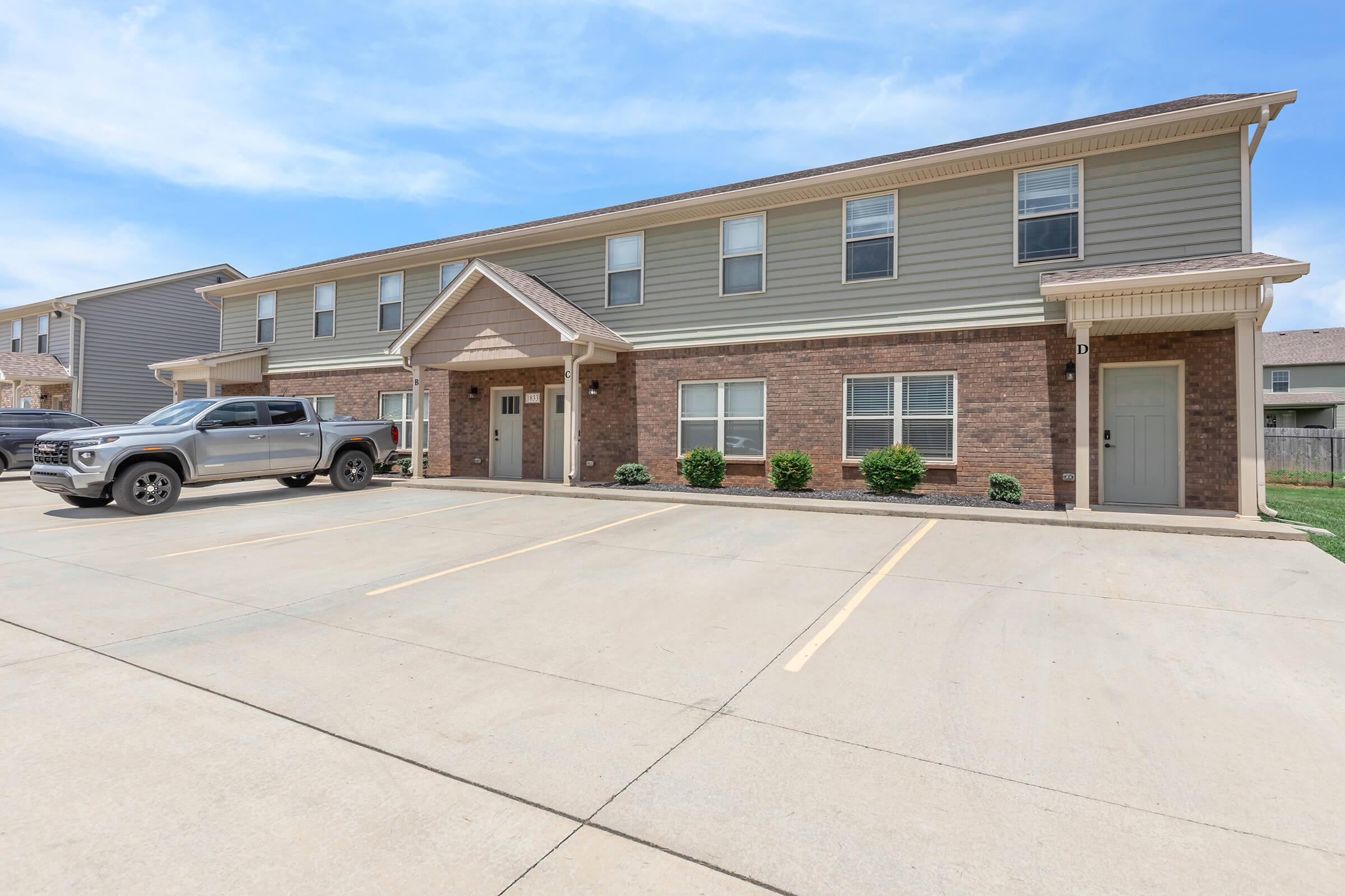 Exterior view of a two-story residential building featuring a mix of brick and siding. A grey pickup truck is parked in front, with well-maintained landscaping and a concrete driveway. The sky is clear and blue, creating a bright atmosphere around the property.