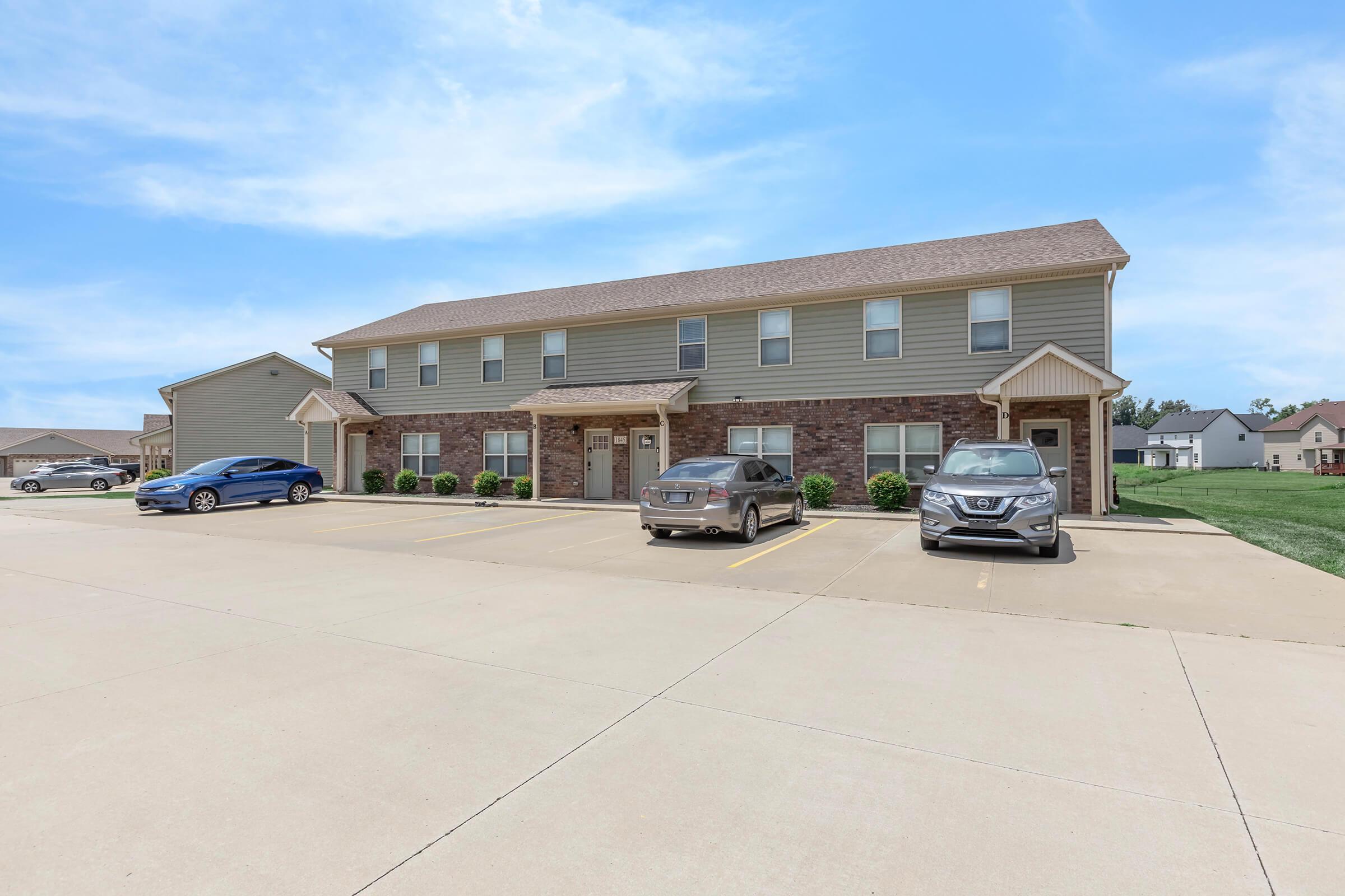 Two-story residential building with a brown brick and light gray siding exterior. Multiple parking spaces are visible in front, with three cars parked. The area is well-maintained, featuring green grass and nearby homes in the background under a blue sky with some clouds.