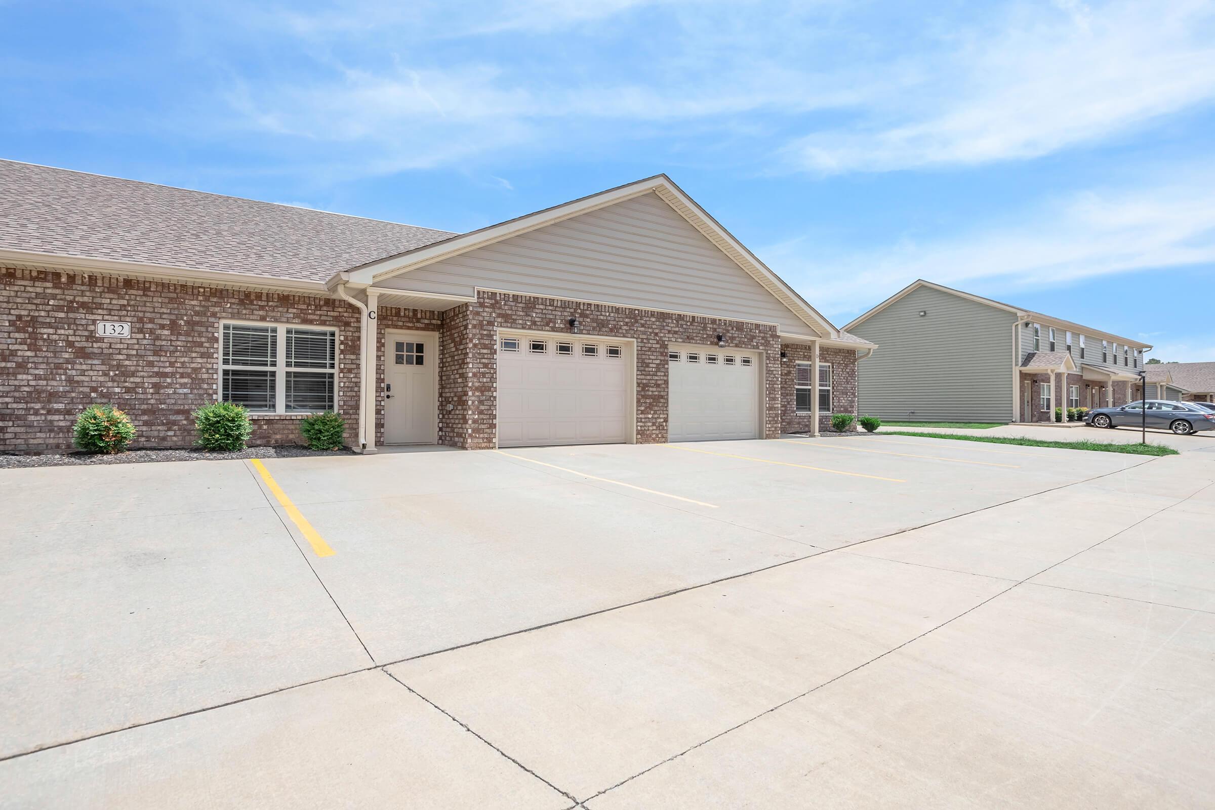 A photo of a residential building featuring brick exterior and a beige façade. The image shows a driveway with two garage doors, surrounded by neatly trimmed shrubs. In the background, another similar building is visible under a clear blue sky.