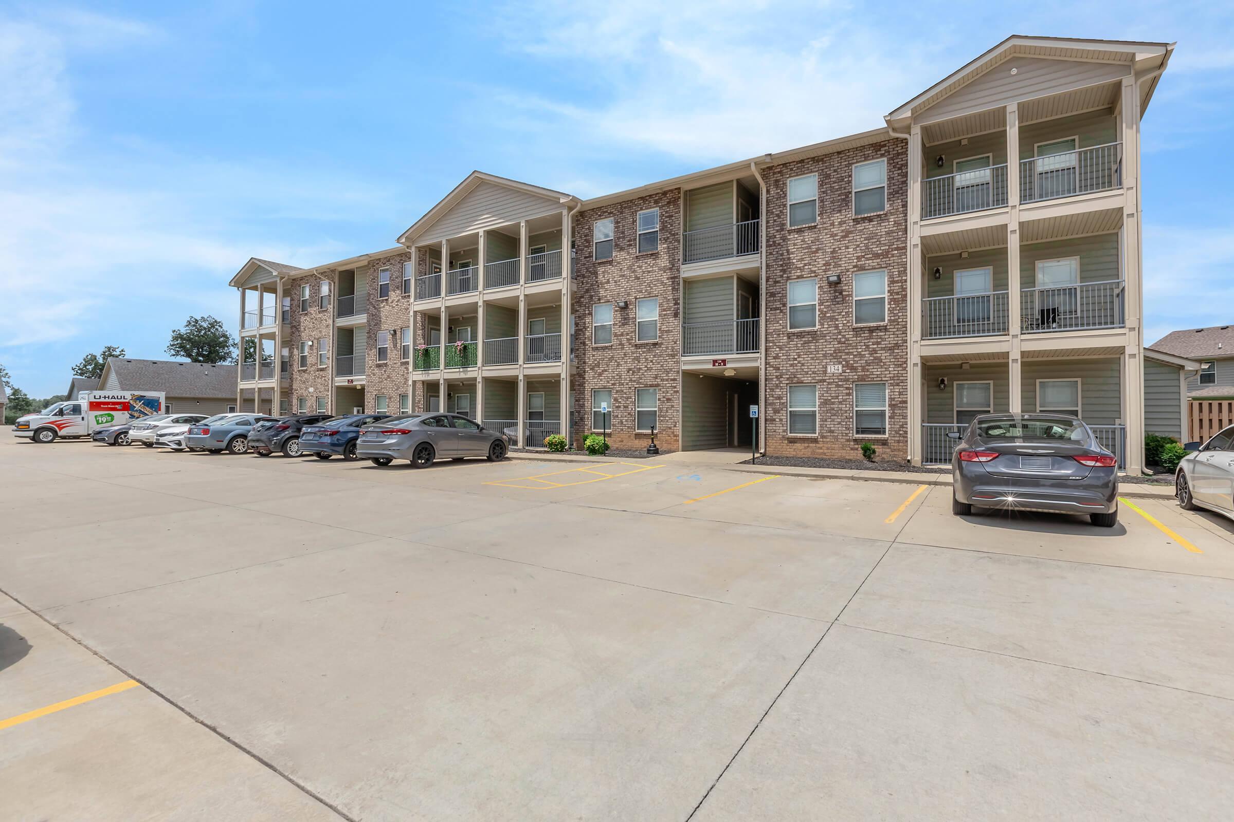 A modern multi-unit apartment building made of brick with multiple balconies. The foreground features a parking lot with several cars parked in designated spaces. The sky is clear and blue, indicating a sunny day.