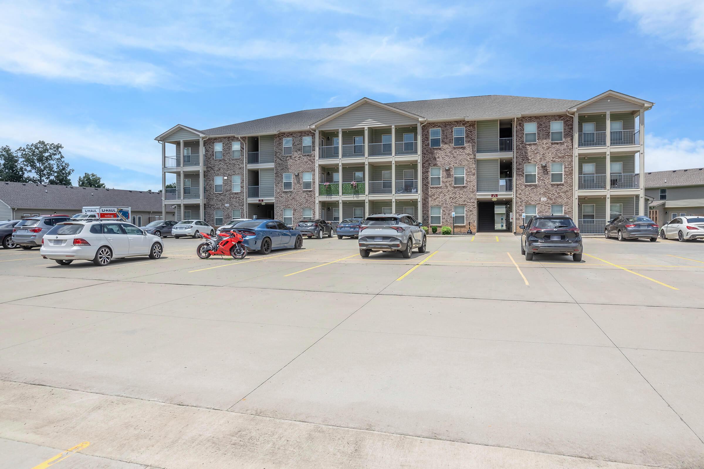 A multi-story apartment building made of brick and siding, featuring several balconies. There is a parking lot in front with various parked cars, including a red motorcycle. The sky is clear and blue, enhancing the building's modern appearance and surrounding greenery.