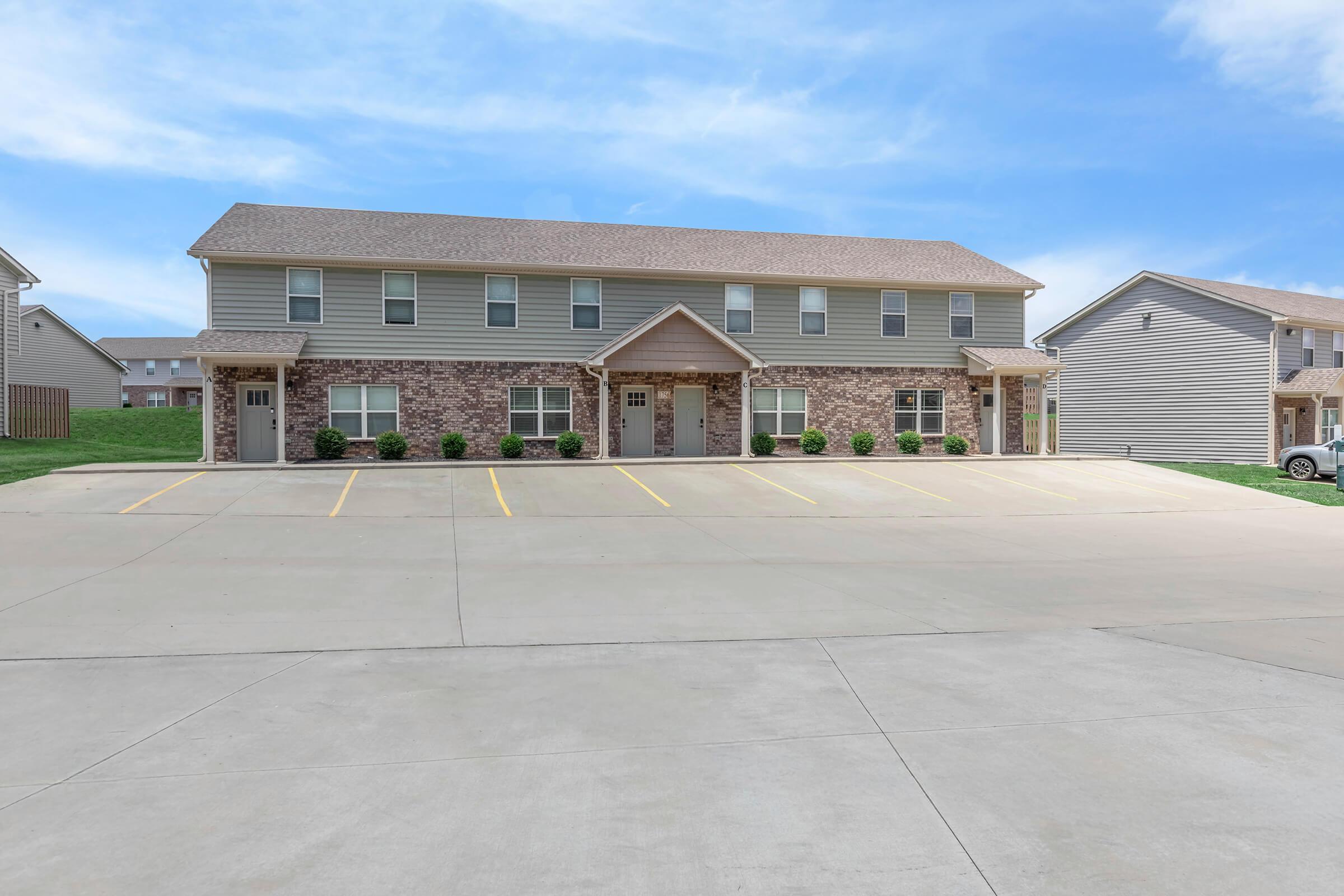 Two-story residential building with a brick and siding exterior, featuring a central entrance. It has several parked vehicles and a spacious concrete lot with marked parking spaces. The surrounding area includes green grass and another building in the background, under a clear blue sky.