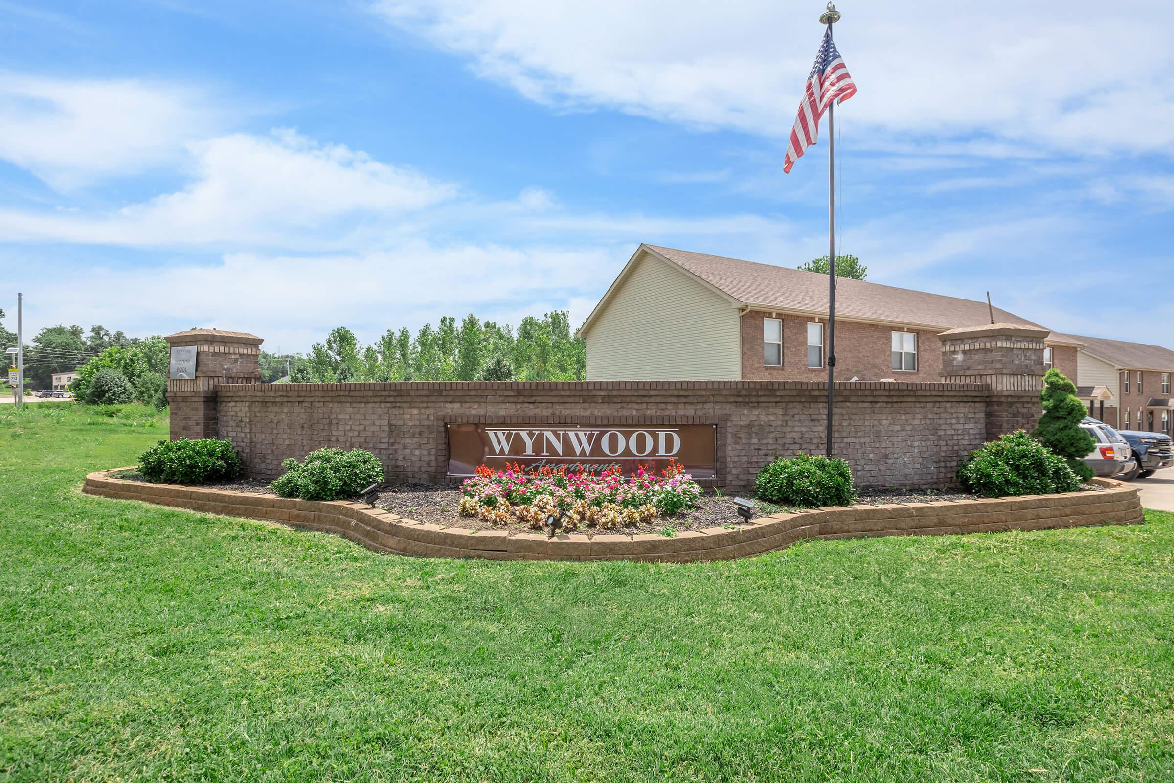 A landscaped entrance sign for Wynwood, featuring colorful flower beds and a flag flying in the background. The setting is a sunny day with a clear blue sky, and there are residential buildings visible in the distance. The area is grassy and well-maintained.