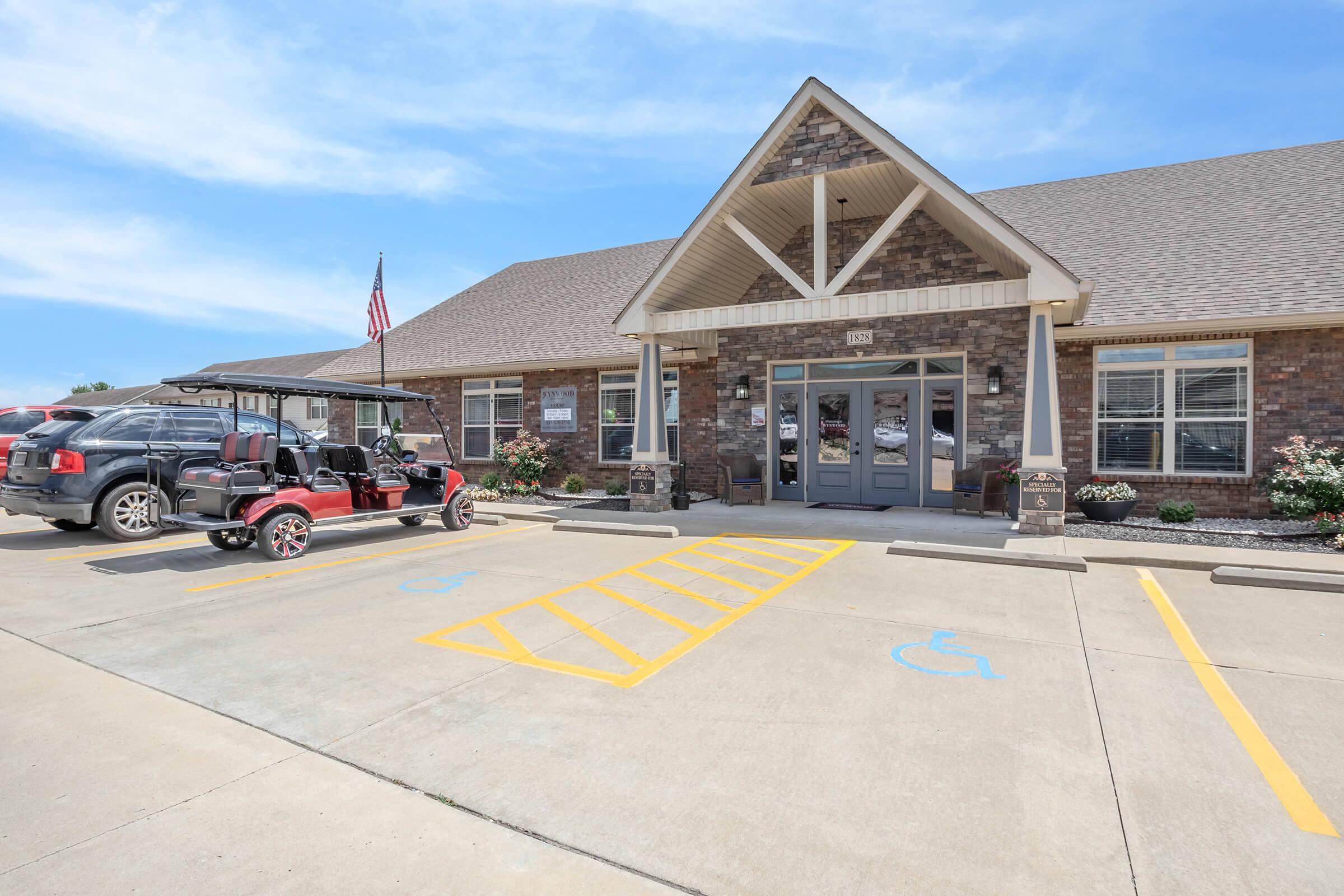 A brick building with a steep, gabled roof houses a welcoming entrance. In front, a parking lot features marked spaces, including handicap spots. A golf cart is parked nearby, and an American flag is visible on the building. Flower beds add greenery to the scene, and the sky is clear and blue.