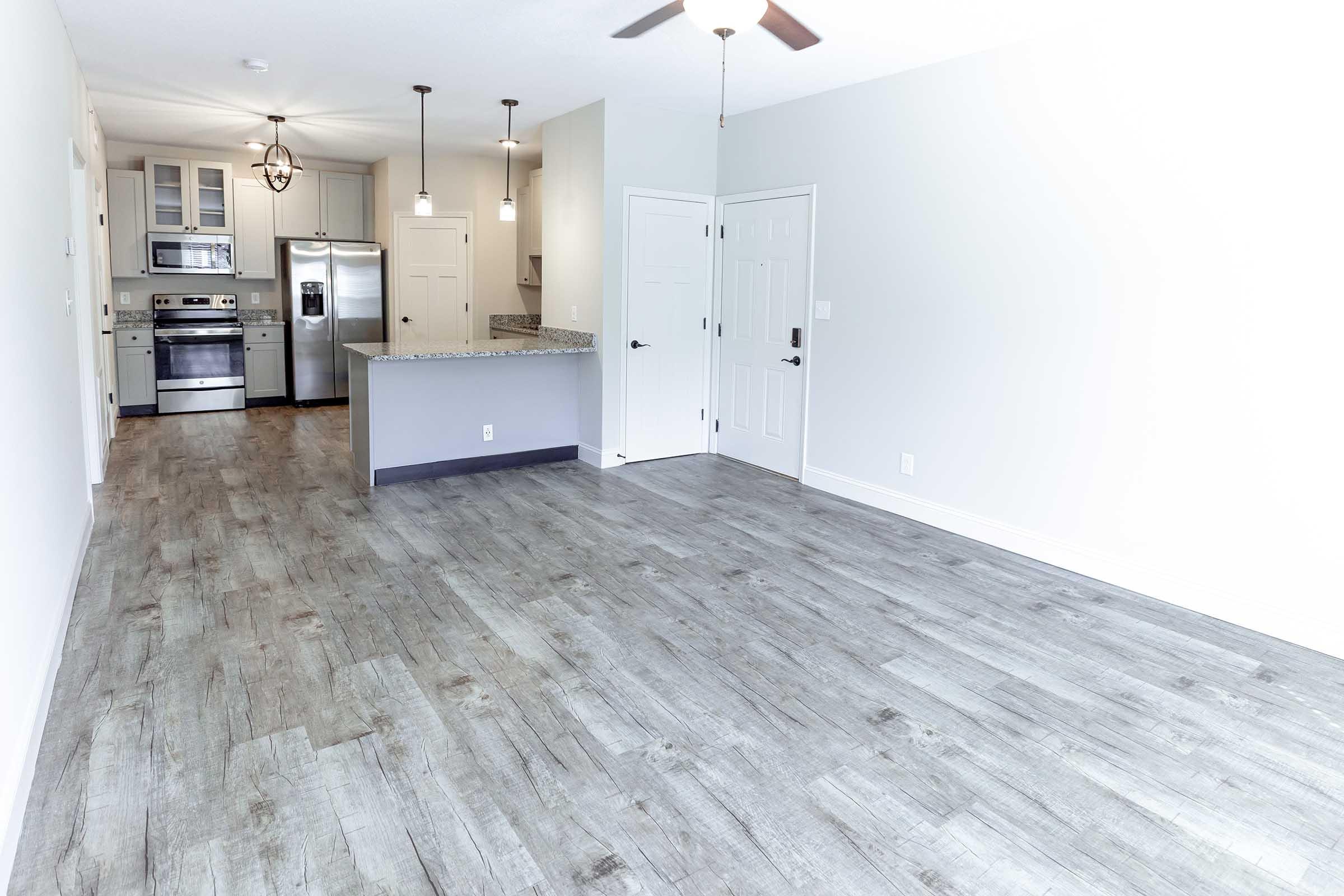 Bright, modern living space featuring an open layout. The room has gray laminate flooring, a kitchen with stainless steel appliances, and a curved breakfast bar. It includes pendant lighting and a ceiling fan, with light-colored walls creating a spacious atmosphere.