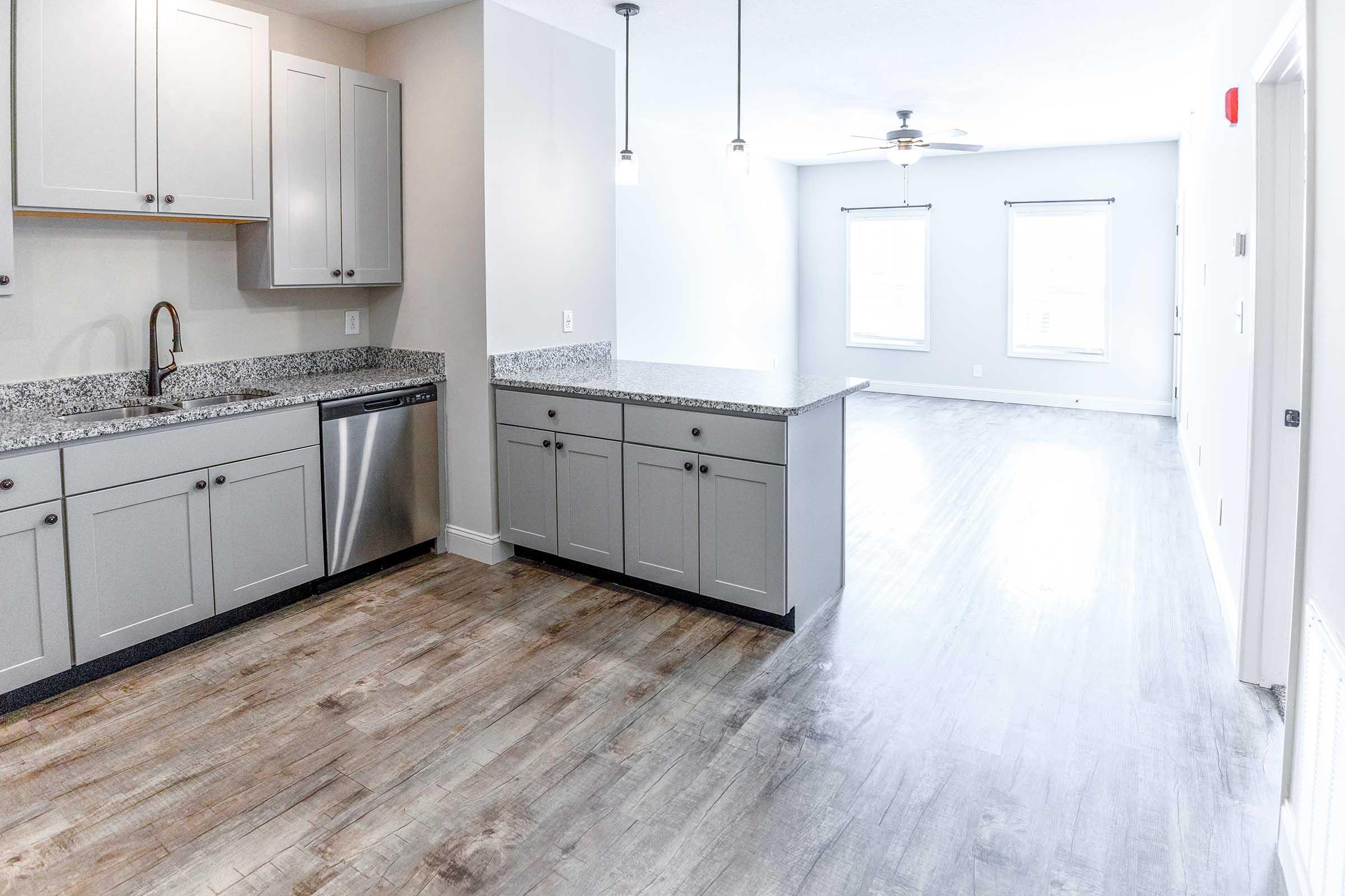 A modern kitchen and living area featuring gray cabinetry, a granite countertop, stainless steel appliances, and a bright, open layout. The kitchen connects to a spacious living room with ceiling fans and large windows overlooking the light-colored flooring.