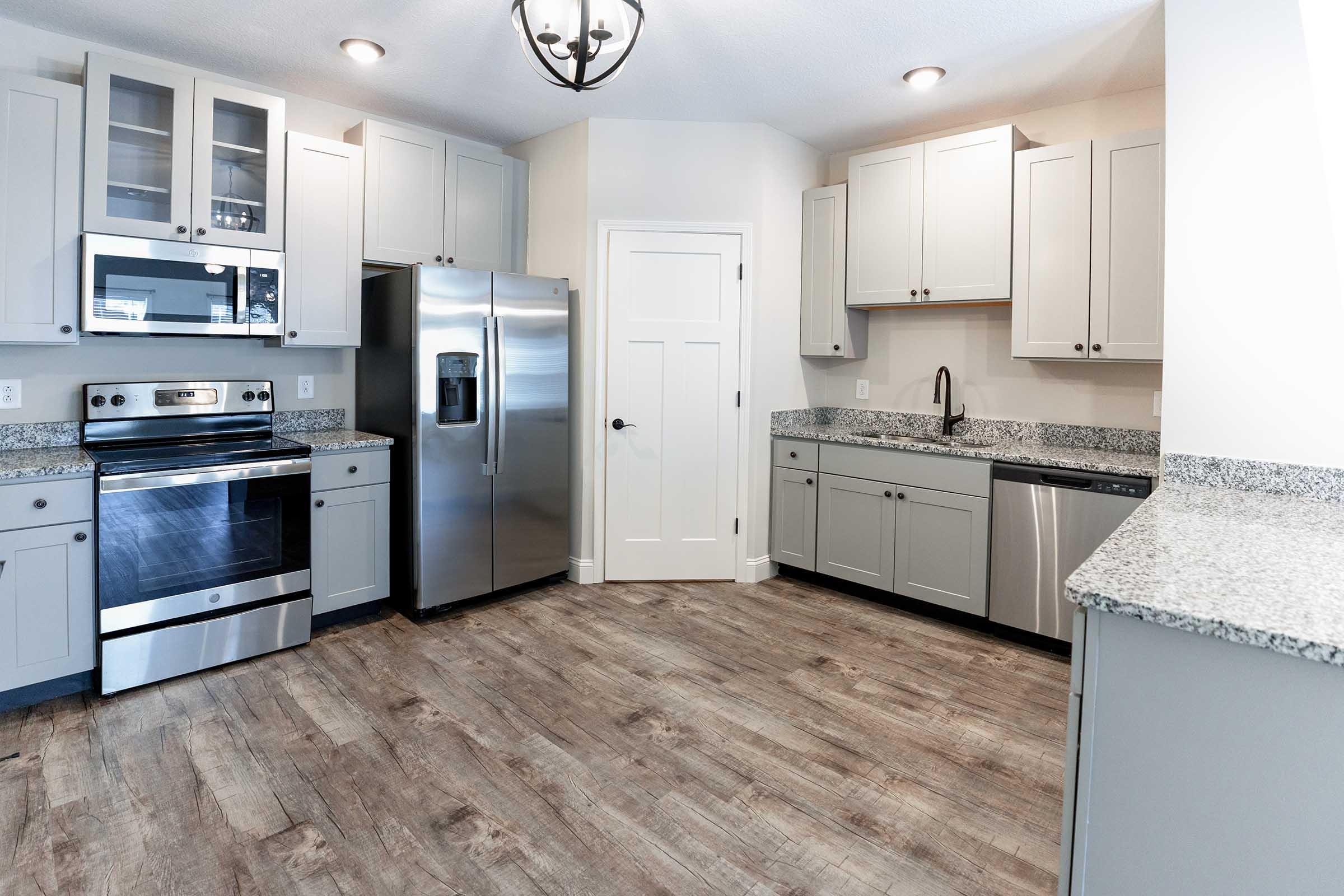 Modern kitchen featuring light gray cabinetry, stainless steel appliances including a refrigerator, oven, and dishwasher, and a spacious countertop with gray speckled granite. The floor has a wood-like finish, and there is ample natural light coming from a nearby window.