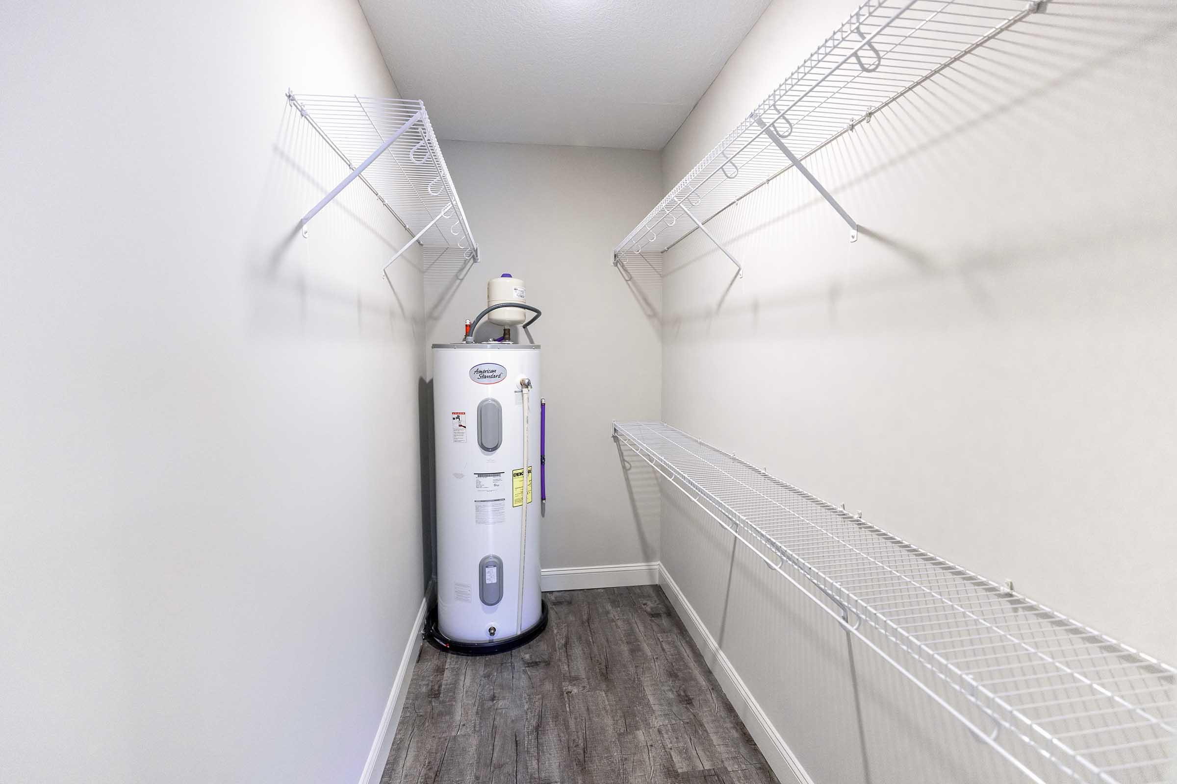 A spacious utility closet with a cylindrical water heater on the left, surrounded by light-colored walls. Two white wire shelving units are installed on the opposite walls, providing ample storage space. The floor features a modern wood-like finish, enhancing the room's overall aesthetic.