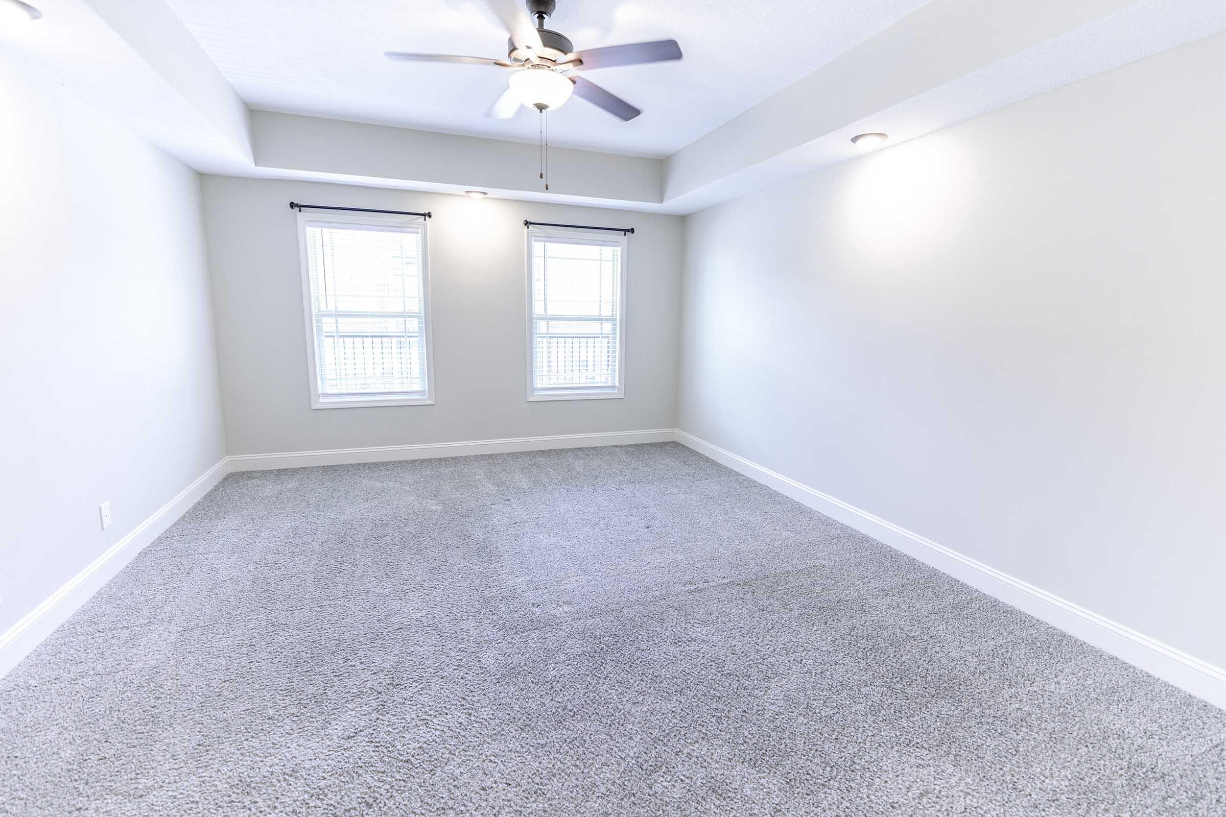 A well-lit, empty room featuring light gray walls, a light-colored carpet, two windows with blinds, and a ceiling fan. The space is spacious and bare, suitable for various uses or décor.