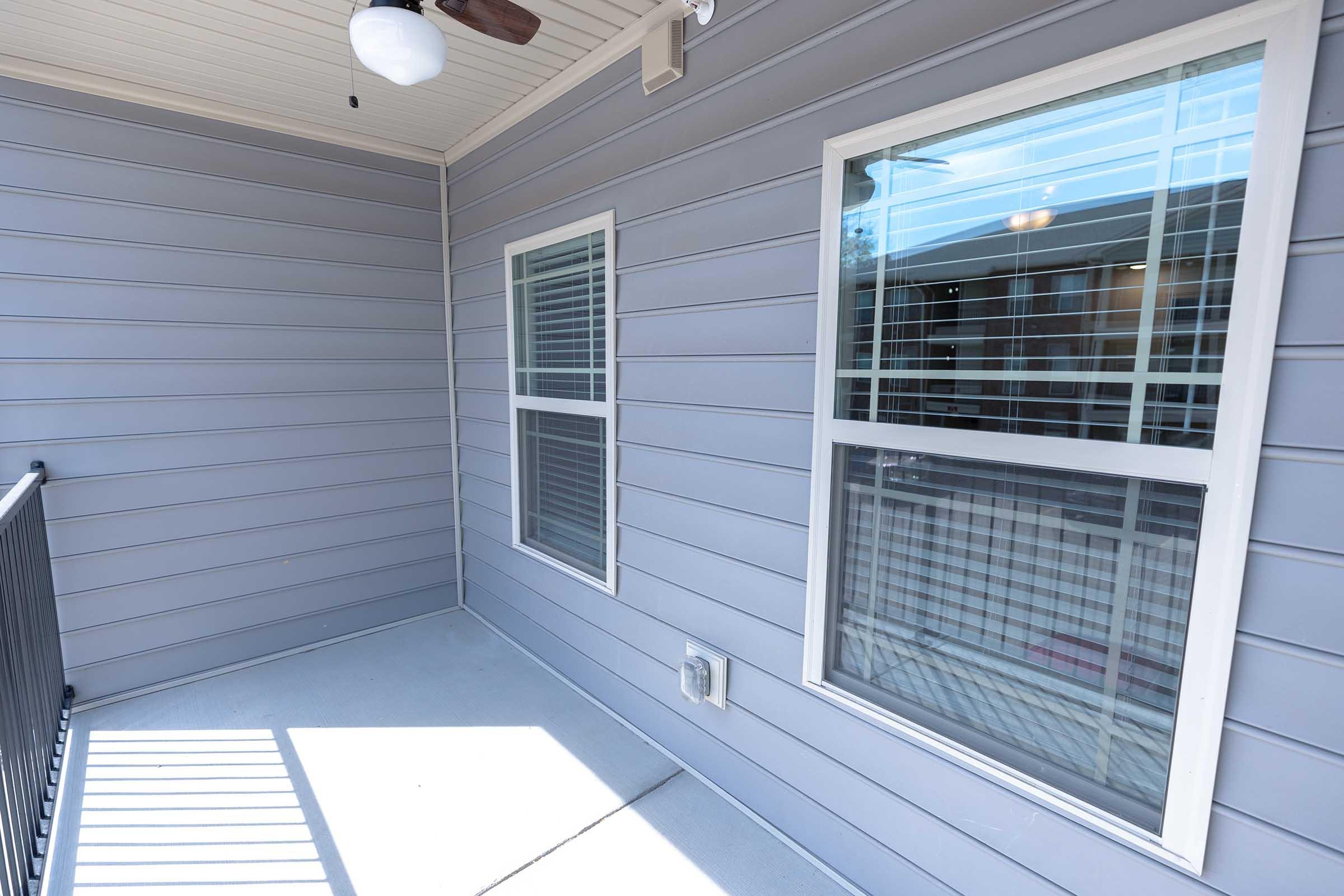 A small covered balcony with grey siding and two windows. Sunlight casts shadows on the concrete floor. The area is clean and well-maintained, with a ceiling fan fixture visible overhead. The background shows reflections from the opposite side.