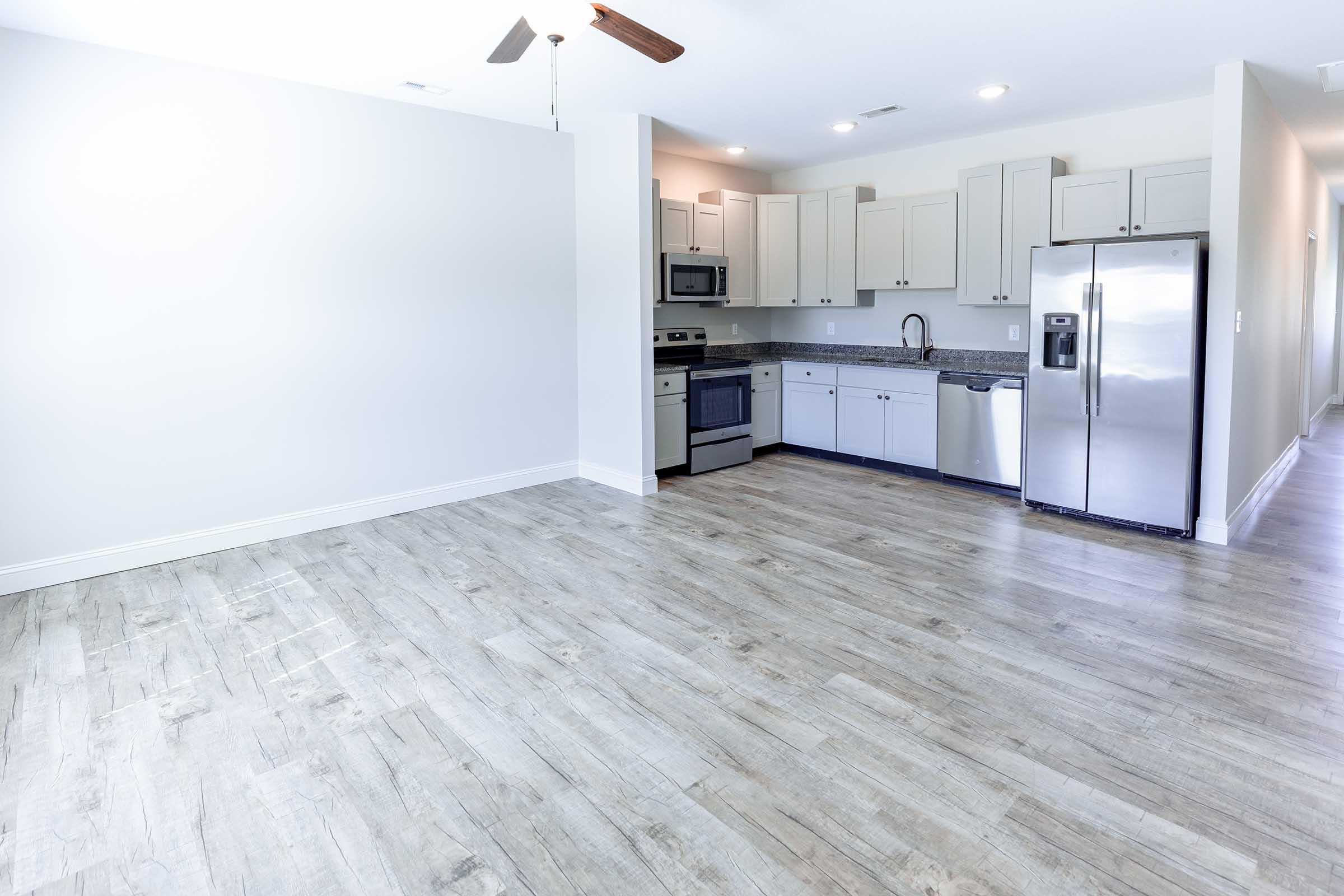 Modern kitchen and living area with gray wooden flooring. The kitchen features light cabinets, stainless steel appliances, and granite countertops. A ceiling fan is visible, and the space is well-lit with natural light, creating a welcoming atmosphere.