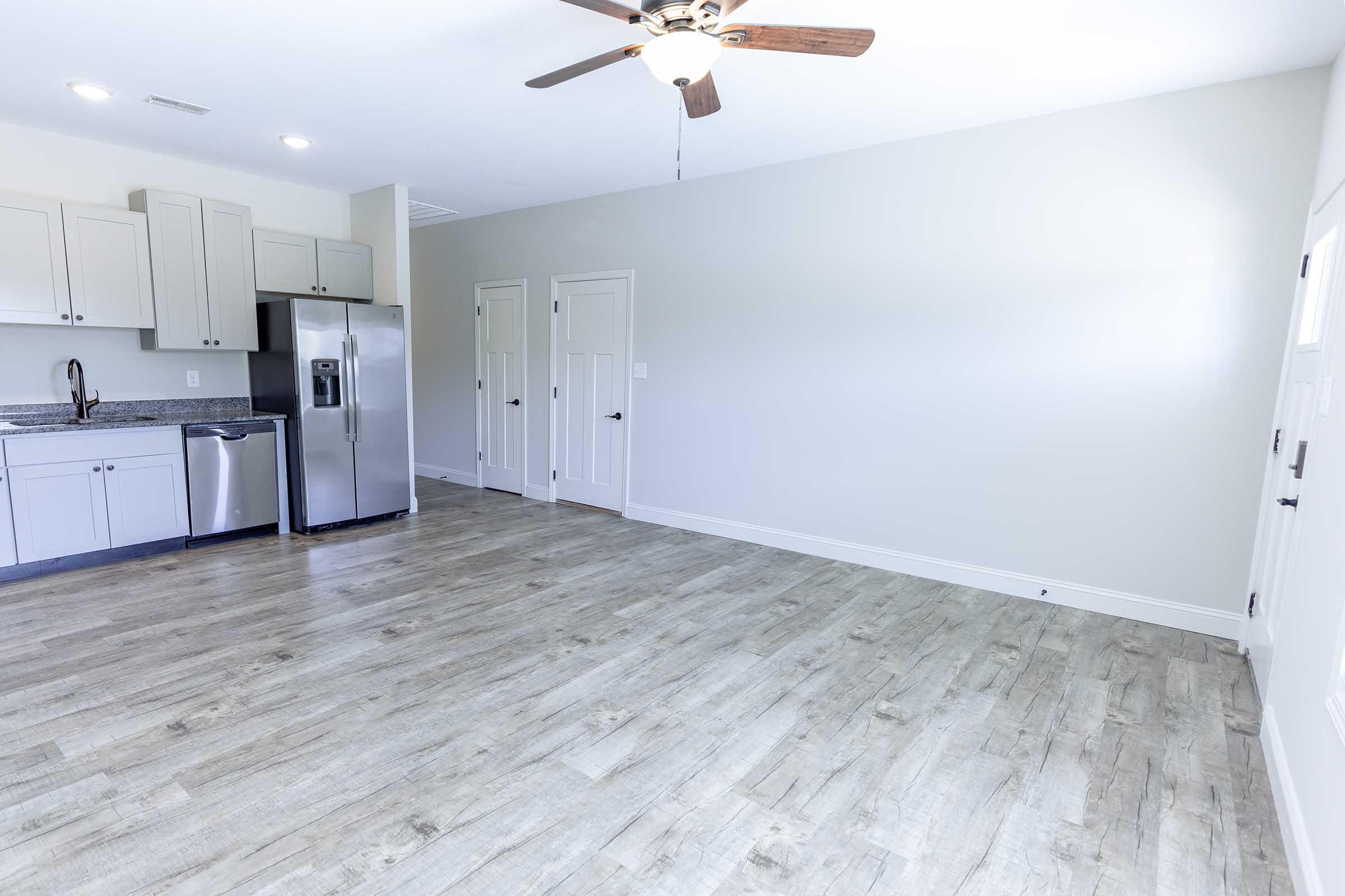 A spacious, well-lit room featuring light gray walls and a ceiling fan. The area includes a modern kitchen with gray cabinets, a stainless steel refrigerator, and a countertop. The flooring is a light wood finish, and there are doors leading to other areas. The room is empty, offering a clean and inviting feel.