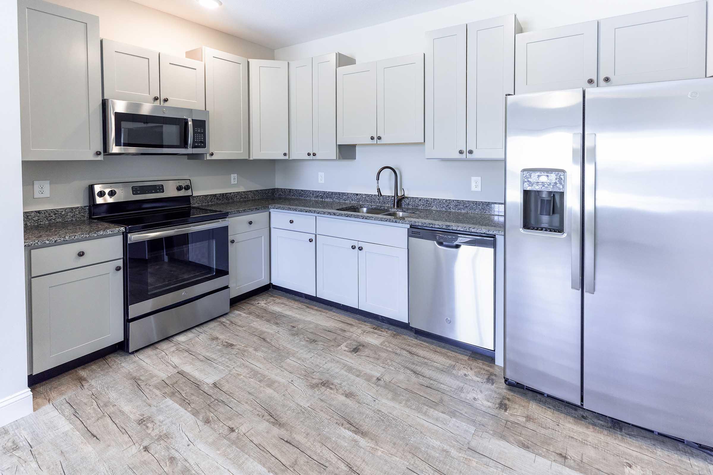 A modern kitchen featuring light gray cabinets, stainless steel appliances including a refrigerator, oven, microwave, and dishwasher, along with a dark granite countertop. The floor is styled with wood-like tiles, creating a contemporary and inviting space.