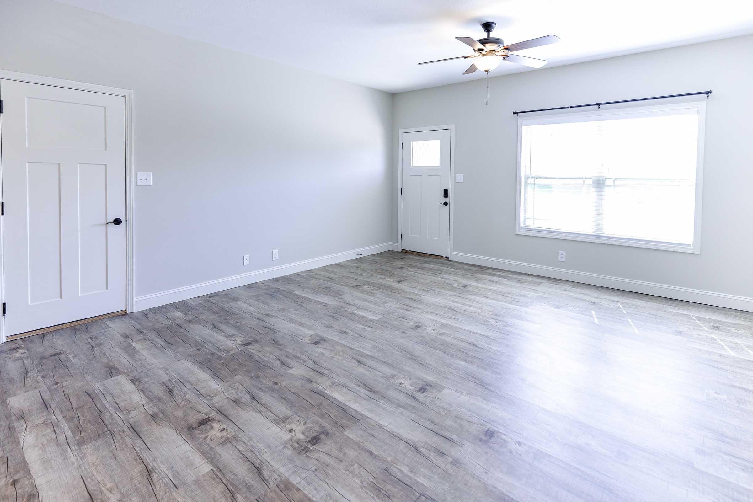 An empty living room featuring light gray walls, a ceiling fan, and large windows with white blinds. The floor is covered with light-colored laminate planks. A closed door is visible on the left, and a front door with a window is on the right. The space is bright and airy, conveying a modern and minimalist aesthetic.