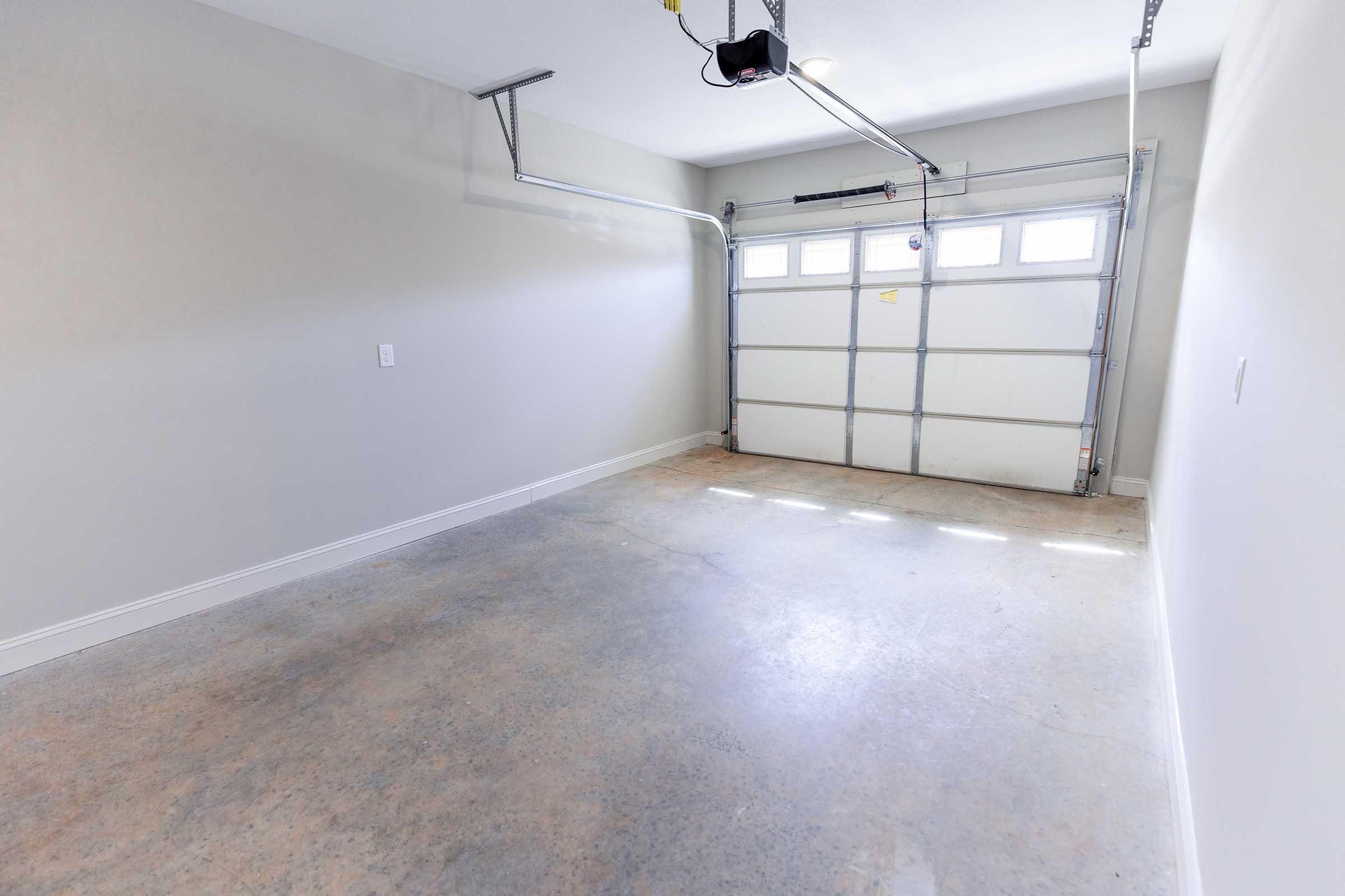 Empty garage space with a concrete floor, featuring a garage door with windows. The walls are painted in a light color, and there are overhead lights. The space is uncluttered, offering a clean and tidy appearance.
