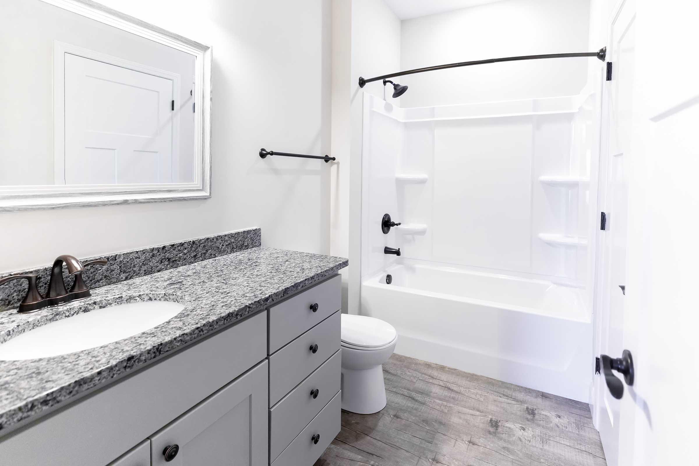 A modern bathroom featuring a white bathtub with shower, a gray vanity with a granite countertop, a sink, a toilet, and black fixtures. The walls are painted white, and there is a large mirror above the sink, with wood-like flooring completing the contemporary design.