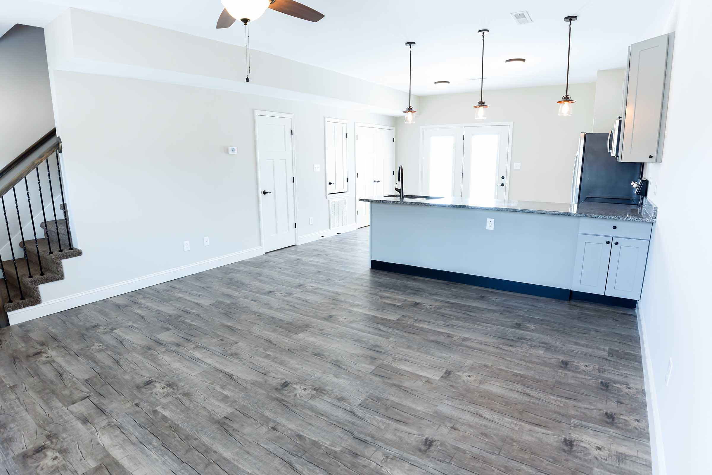A spacious modern living area with light gray walls and wood-like laminate flooring. The kitchen features a breakfast bar with granite countertops, pendant lighting, and stainless steel appliances. There is a staircase in the background and bright natural light coming from windows and doors.