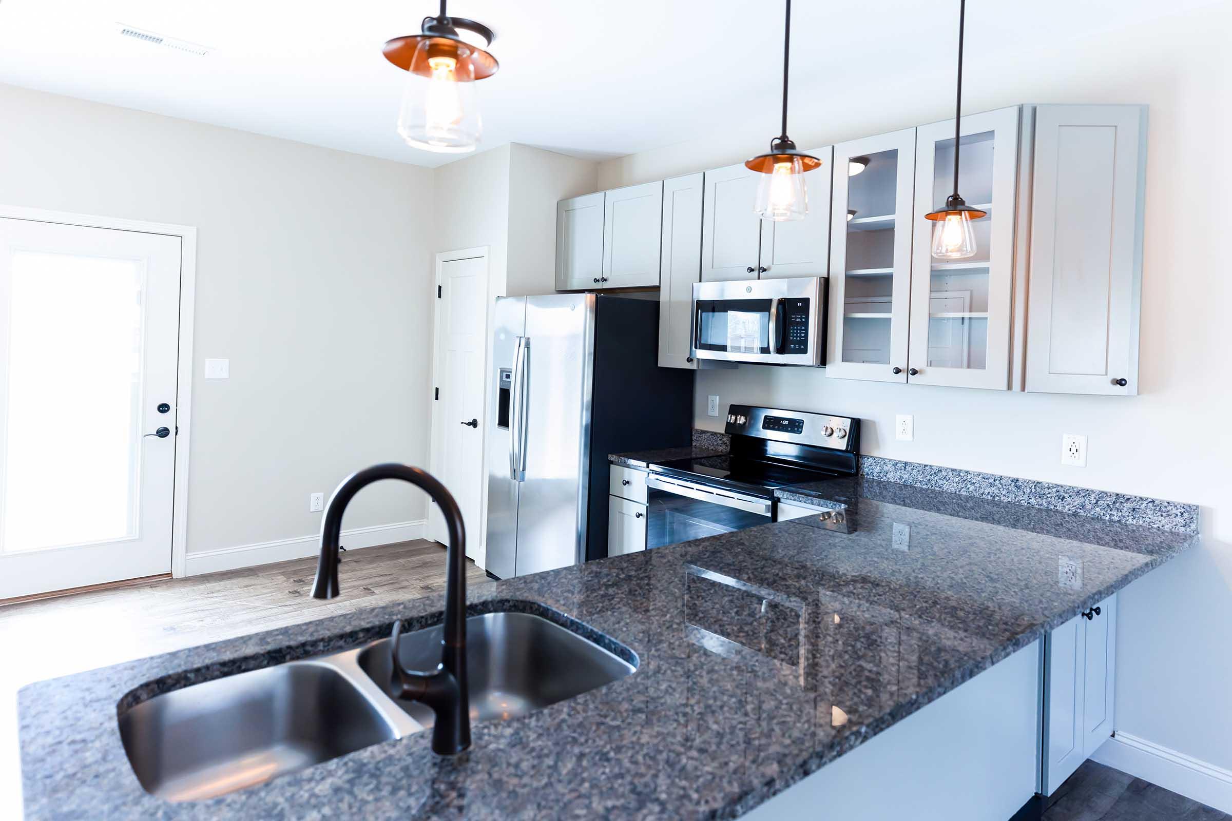 Modern kitchen featuring granite countertops, stainless steel appliances including a refrigerator, microwave, and oven. The space is illuminated by pendant lighting, and there is a double sink with a sleek black faucet. Light-colored cabinets and a door leading outside complement the clean and contemporary design.