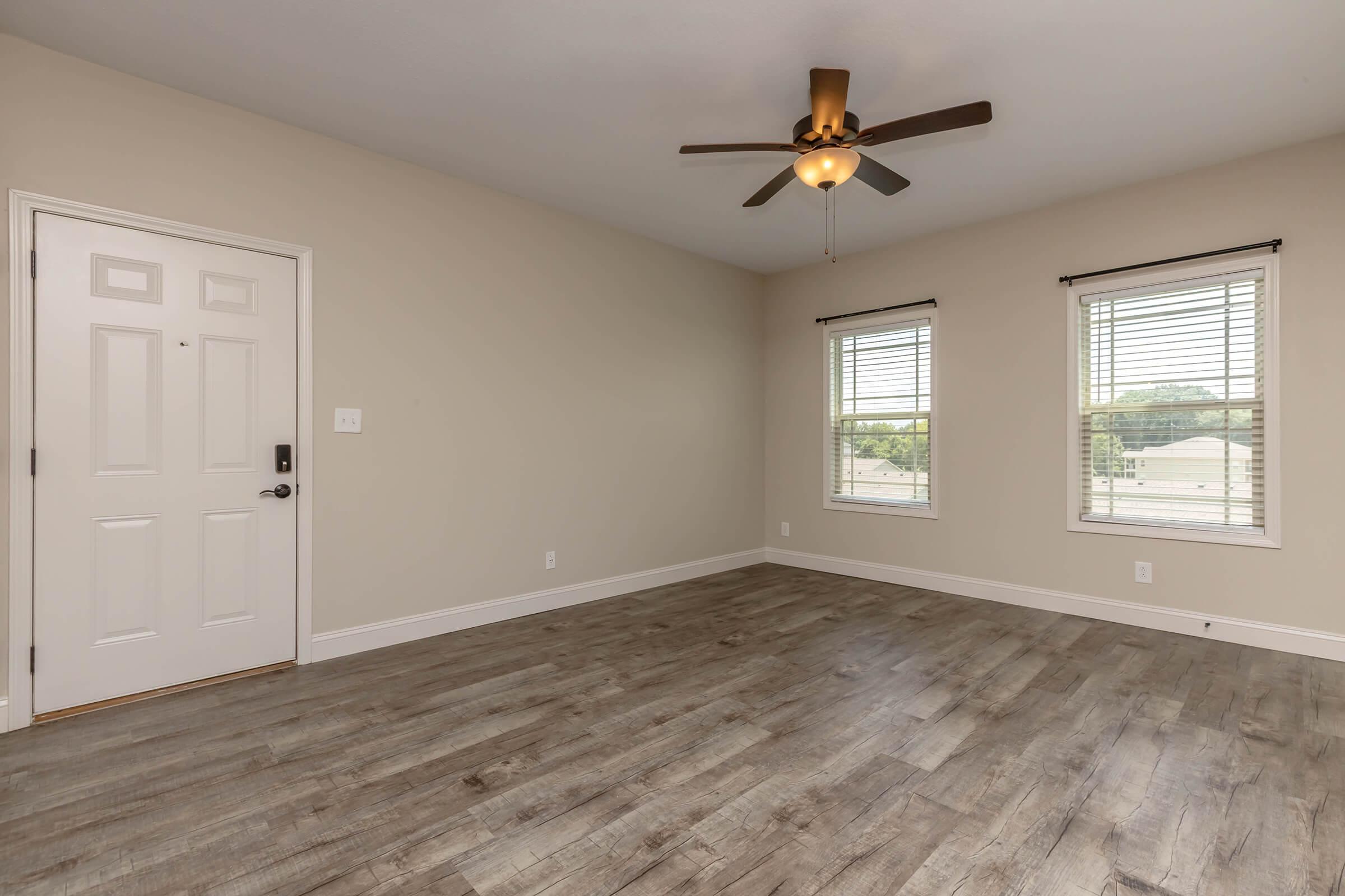 Empty room with light beige walls and wooden flooring. A ceiling fan is installed above, and there are two windows with blinds on the right side. The door is on the left, featuring a simple design. Natural light brightens the space, creating a clean and airy atmosphere.