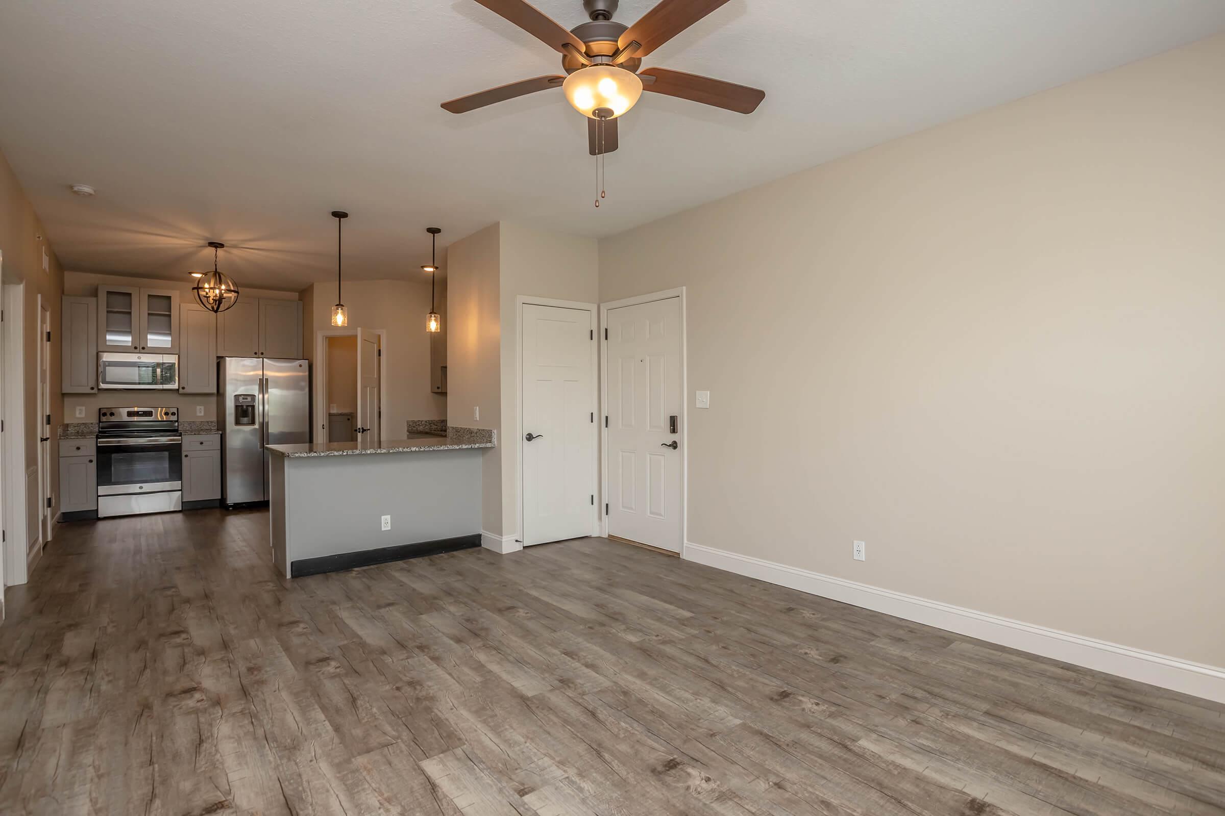 Spacious living area with light wood-like flooring, featuring a ceiling fan. In the background, a modern kitchen is visible with stainless steel appliances and a granite countertop. Two pendant lights hang above the kitchen island, and there are two closed doors leading to other rooms. The walls are painted a neutral beige.