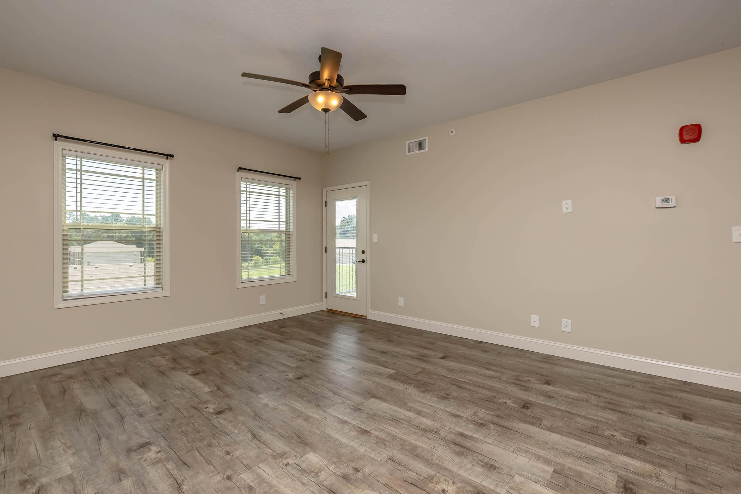 Spacious, empty room with wood-like flooring, a ceiling fan, and two windows with blinds. A door leads to a balcony, and there are wall controls and a smoke alarm visible. The walls are painted a neutral color, creating a bright and inviting atmosphere.