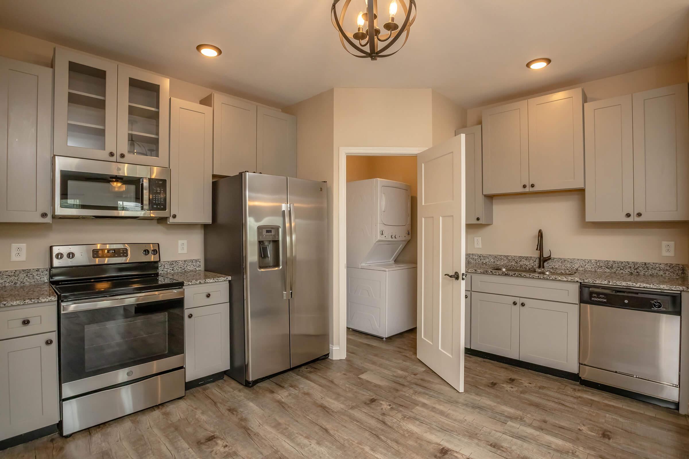 Modern kitchen featuring stainless steel appliances, including an oven, microwave, and refrigerator. The cabinets are light gray, complemented by granite countertops. There is a dishwasher beside the sink. A door leads to a laundry area beyond the kitchen, with wood-style flooring throughout.