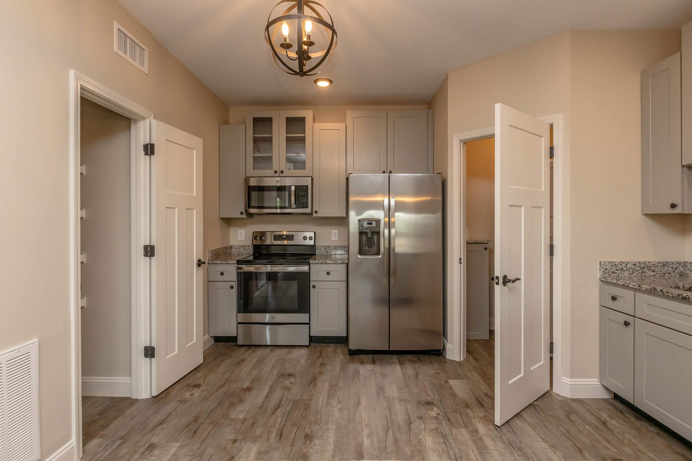 Modern kitchen featuring stainless steel appliances, including a refrigerator and oven, with light gray cabinets and granite countertops. The space has a neutral wall color and a wooden floor. Two open doors provide access to adjacent rooms, and a contemporary chandelier hangs from the ceiling.