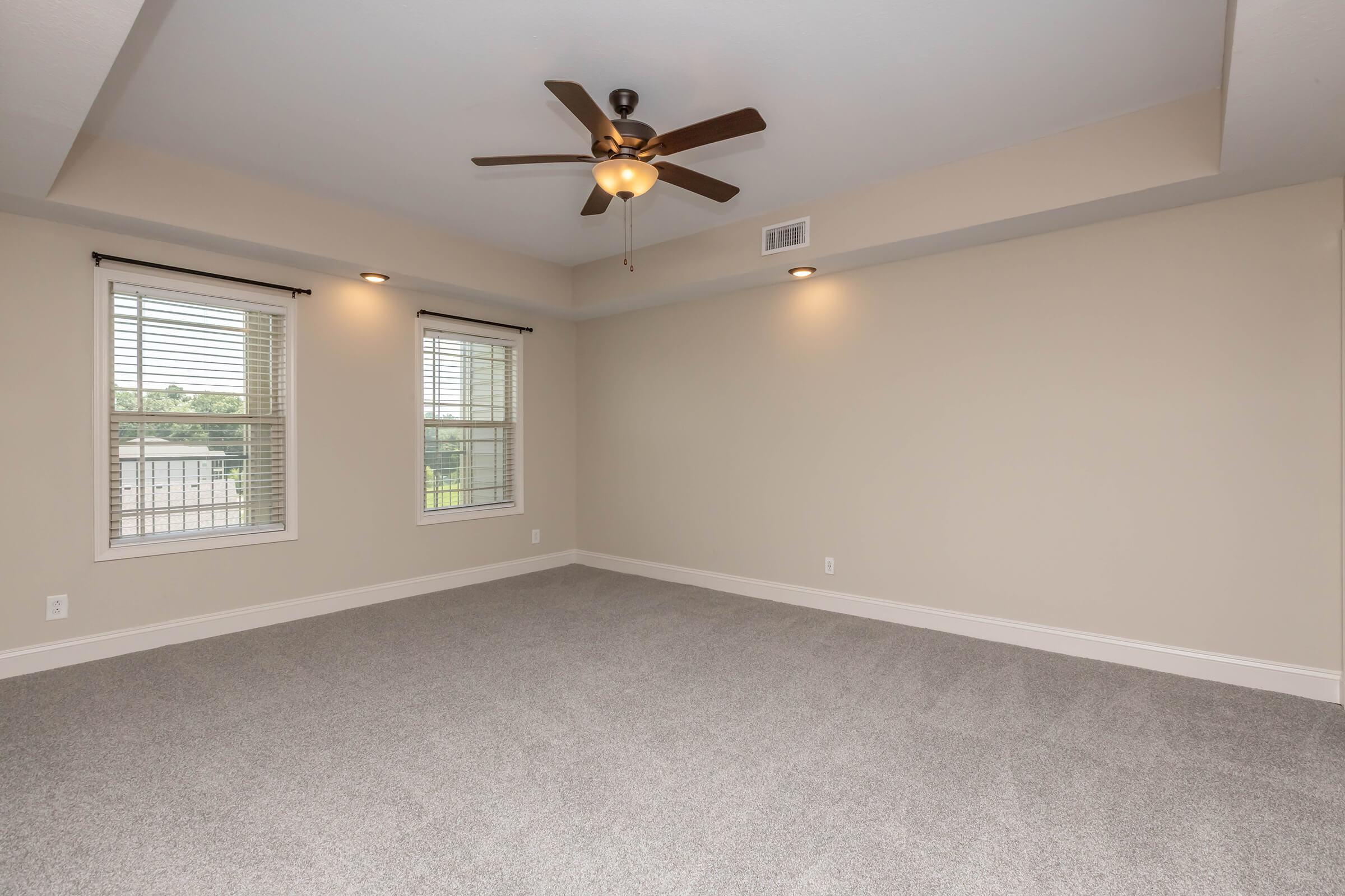 A spacious, well-lit room with light beige walls and new carpet. It features two windows with blinds, allowing natural light to enter, and a ceiling fan mounted above. The room has a simple design with no furniture, creating a blank canvas for decoration.