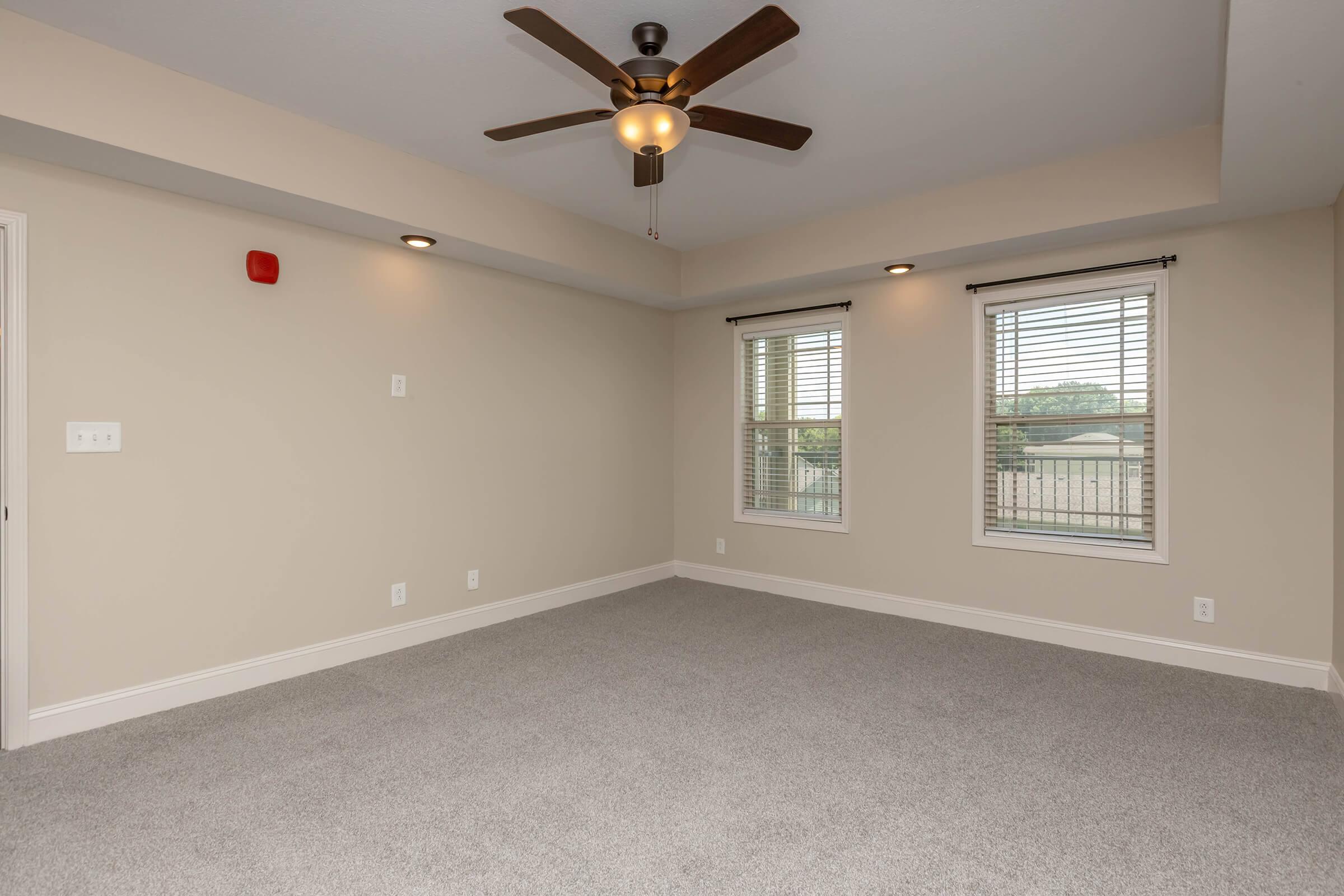 Spacious, empty room with light beige walls and gray carpet. It features large windows with white blinds, a ceiling fan with wooden blades, and minimal lighting fixtures. One wall has a red fire alarm. The room has a bright and airy feel, suitable for various uses such as a living area or bedroom.