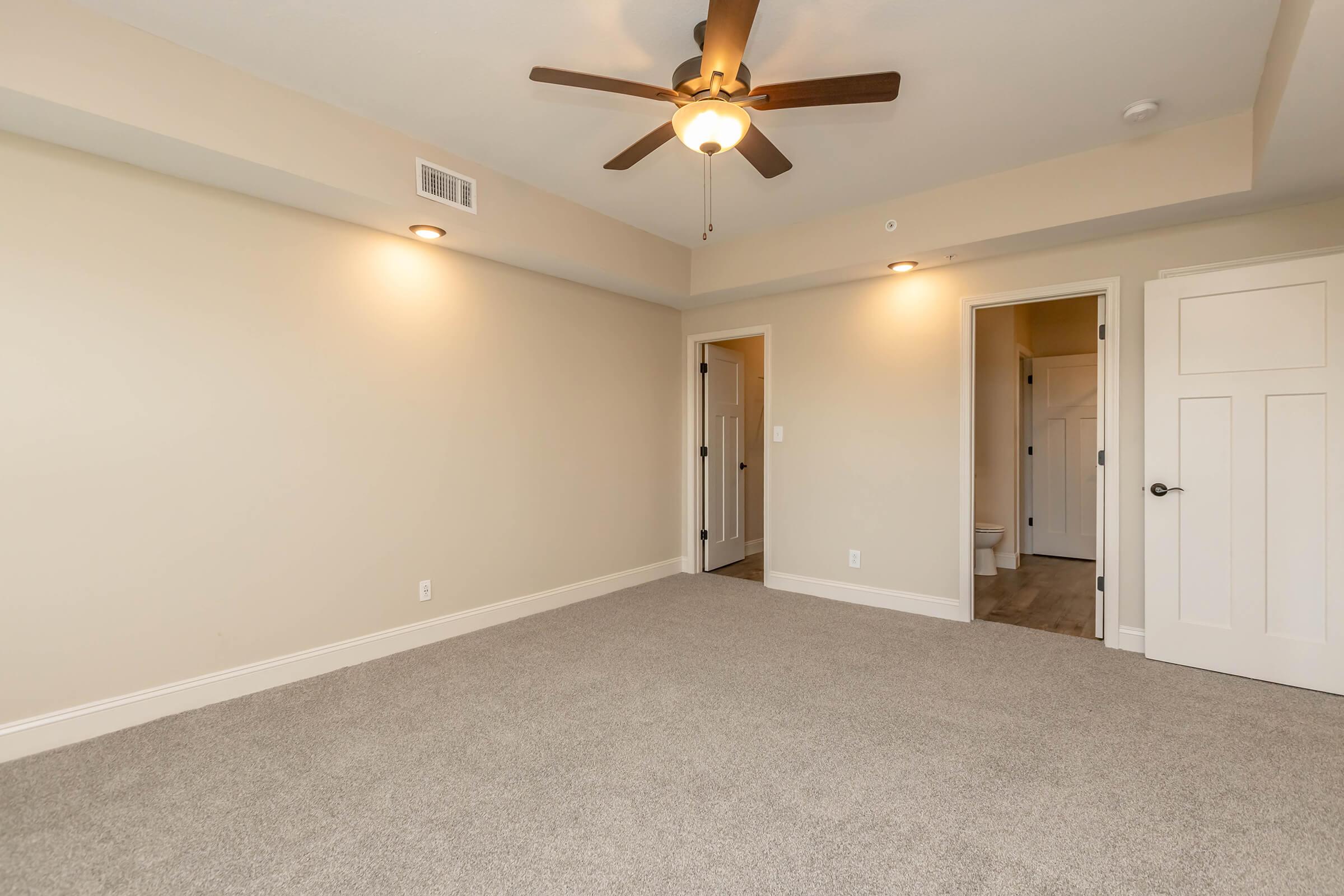 A spacious empty room with light beige walls and a ceiling fan. The floor is covered in carpet and there are two doorways leading to adjacent rooms. Soft lighting fixtures are installed in the ceiling, creating a warm ambiance.