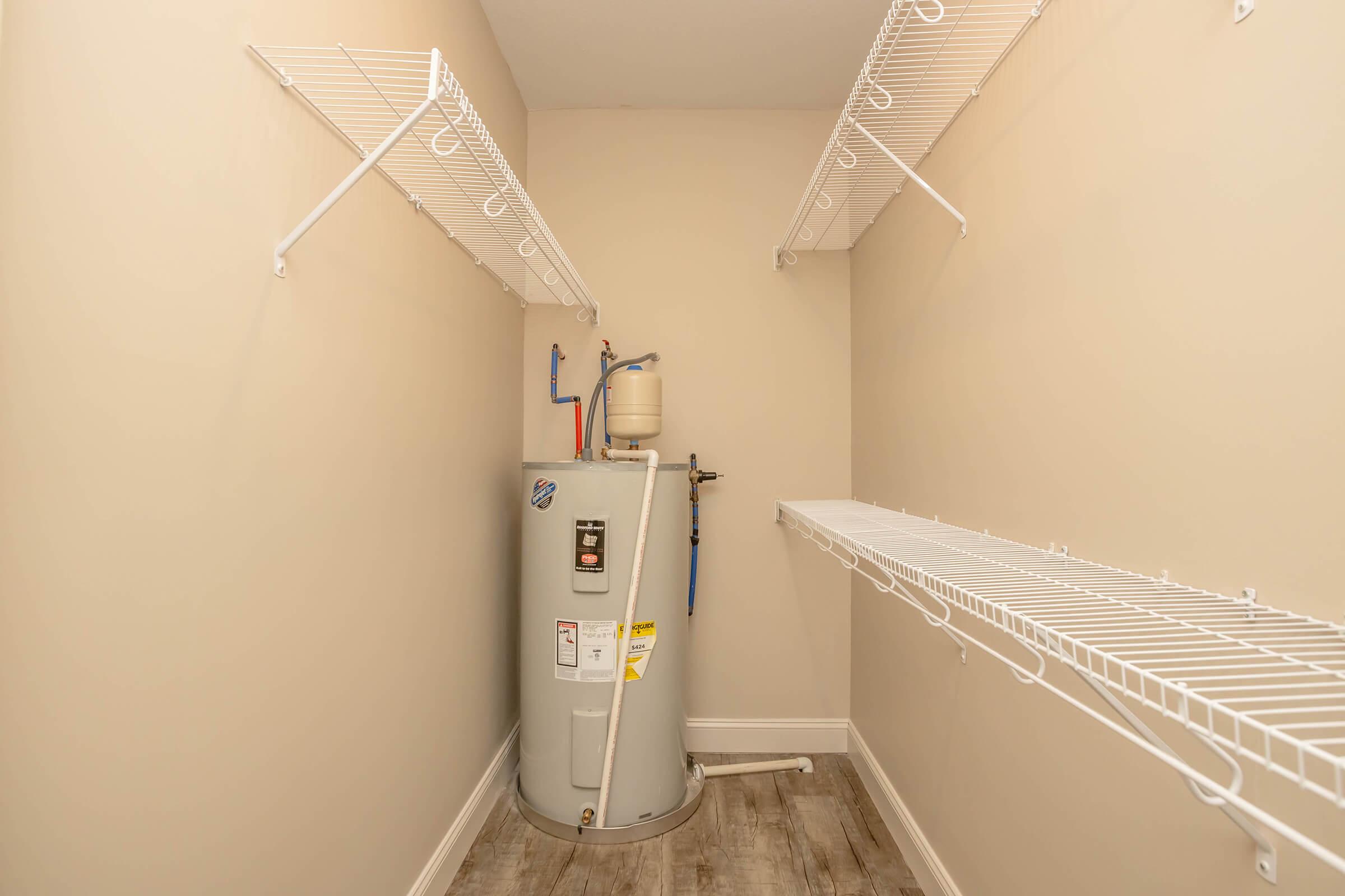 A small utility room with light beige walls and wooden flooring. A grey water heater is positioned against one wall. The space features two white wire shelving units on opposite walls, providing storage options. The room is well-lit and uncluttered, focusing on functionality.