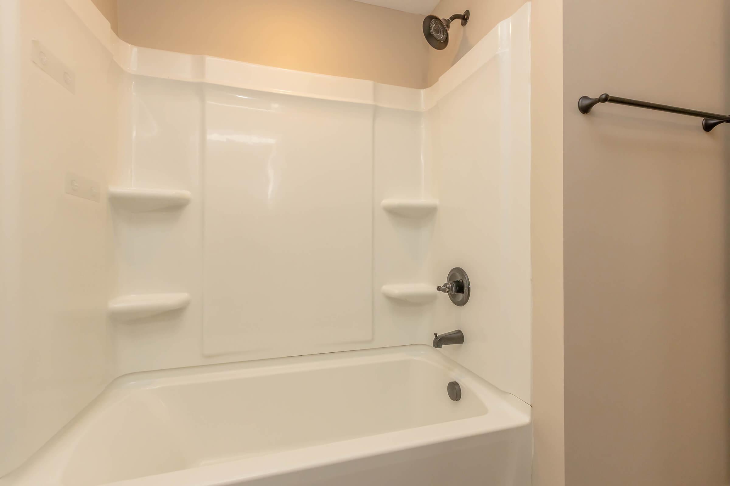 A clean and simple bathroom with a white bathtub and shower combination. The walls are smooth and light-colored, with soap shelves embedded in the shower walls. A chrome faucet and showerhead complete the setup, while a towel bar is visible on the adjacent wall. The overall design is modern and minimalistic.