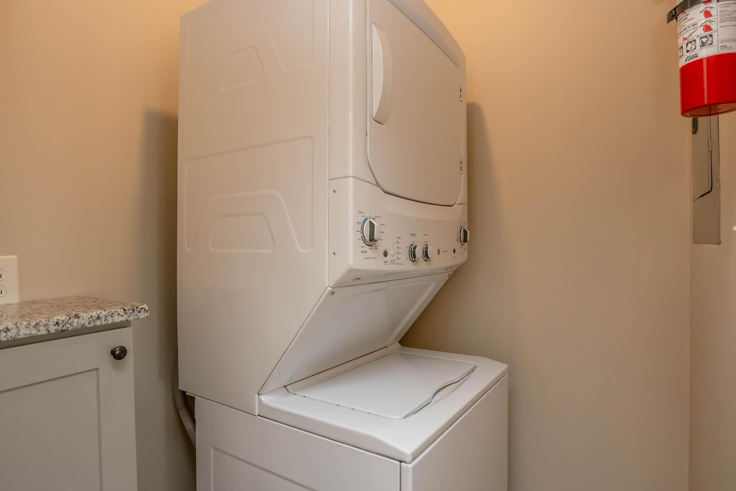 A stacked washer and dryer unit in a laundry room, positioned against a wall with a beige background. A fire extinguisher is mounted nearby, and there is a countertop next to the appliance. The space appears clean and organized.