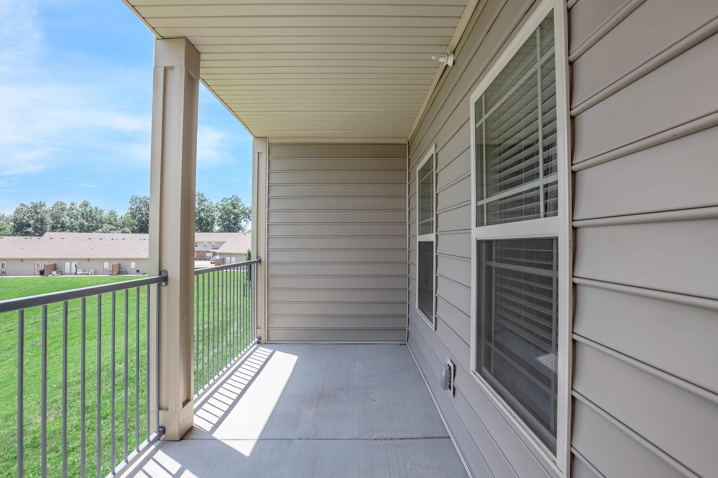 A partially shaded balcony with a light-colored railing and wall. The floor features concrete, and there are large windows showcasing a view of a grassy area and nearby buildings in the background under a clear blue sky.