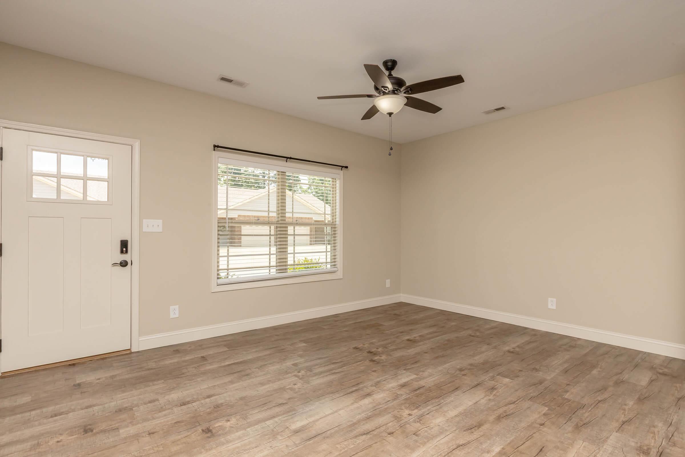 A spacious, empty room featuring light gray walls, a ceiling fan, and wood-like flooring. There's a window with blinds allowing natural light in and a front door on the left side. The room has a clean, minimalist aesthetic, ideal for various furnishings and decorations.