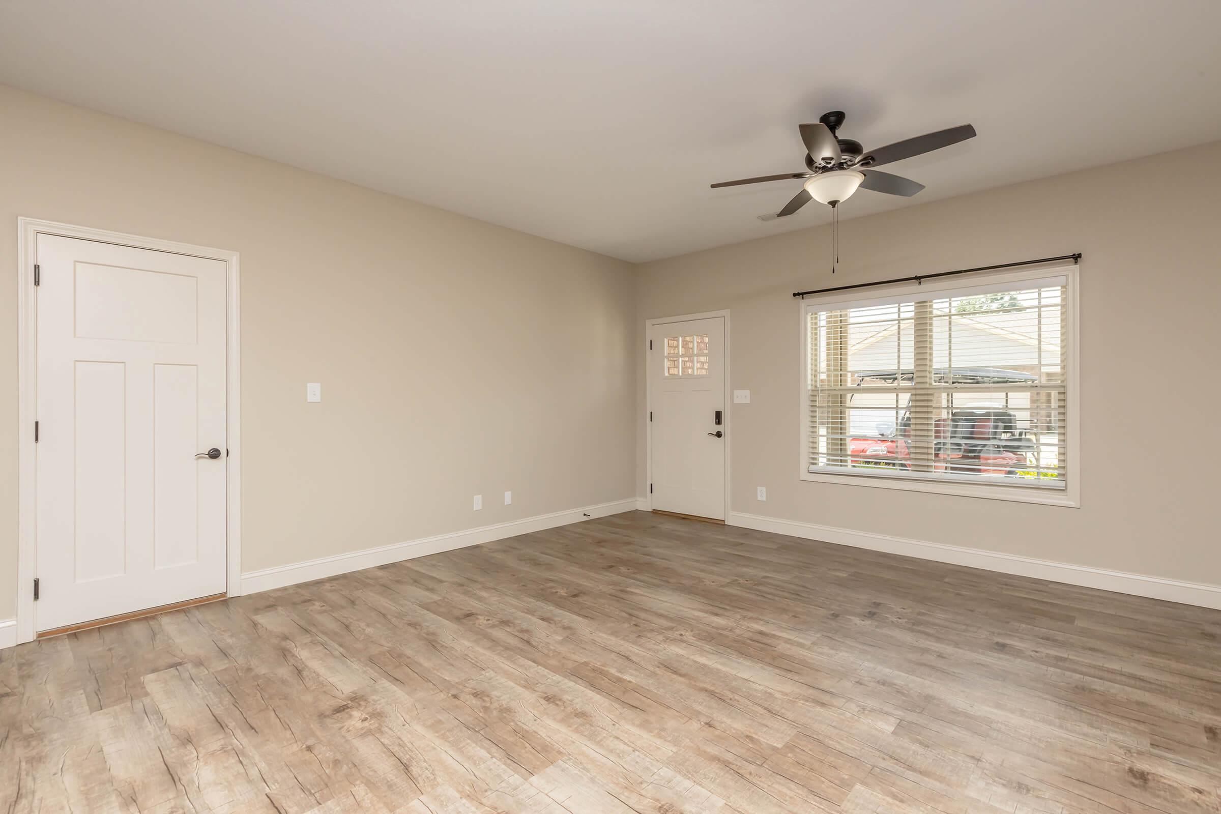 Empty room featuring light beige walls, a ceiling fan, and large window with blinds. The floor has a wood-like finish. A closed door is visible on the left, and there's a window leading outside on the right, adding natural light to the space. The room appears spacious and ready for furnishing.