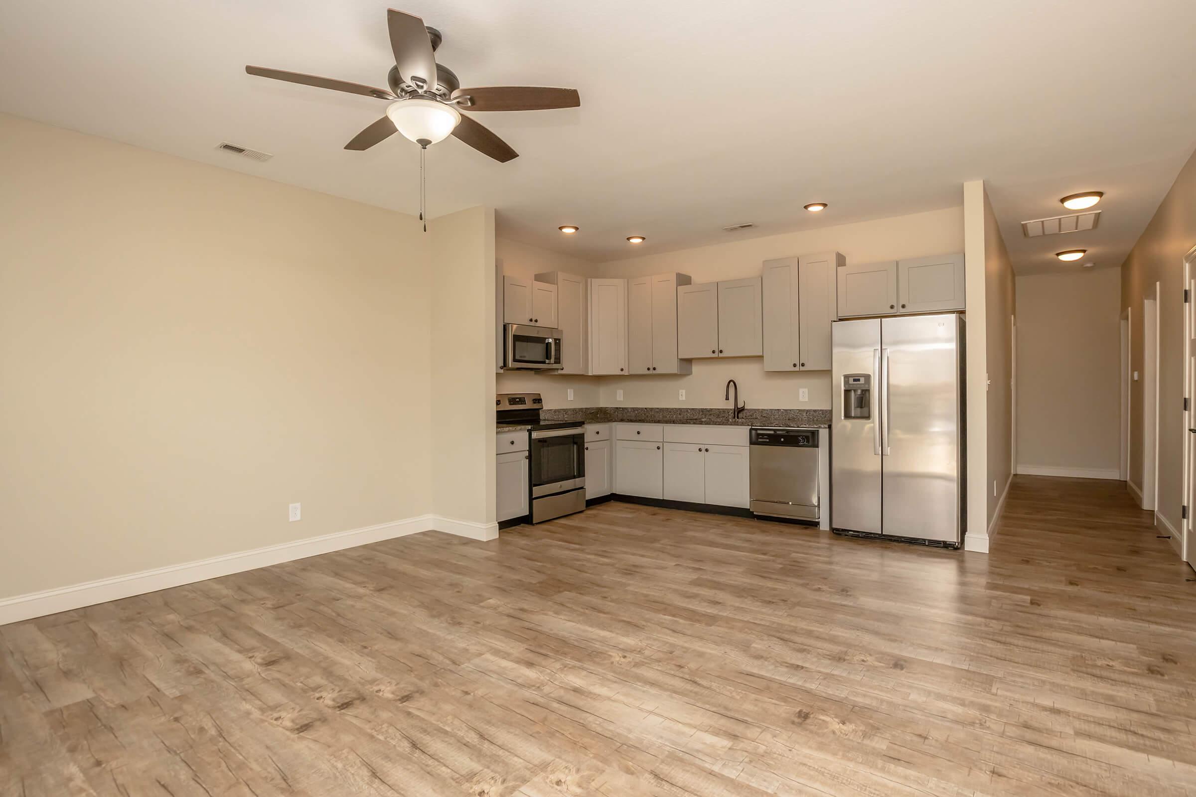 A modern kitchen and living area with light-colored walls, wood-style flooring, and a ceiling fan. The kitchen features stainless steel appliances, a microwave, and an island with bar seating. Adjacent to the kitchen is a hallway leading to closed doors, suggesting access to bedrooms or bathrooms.