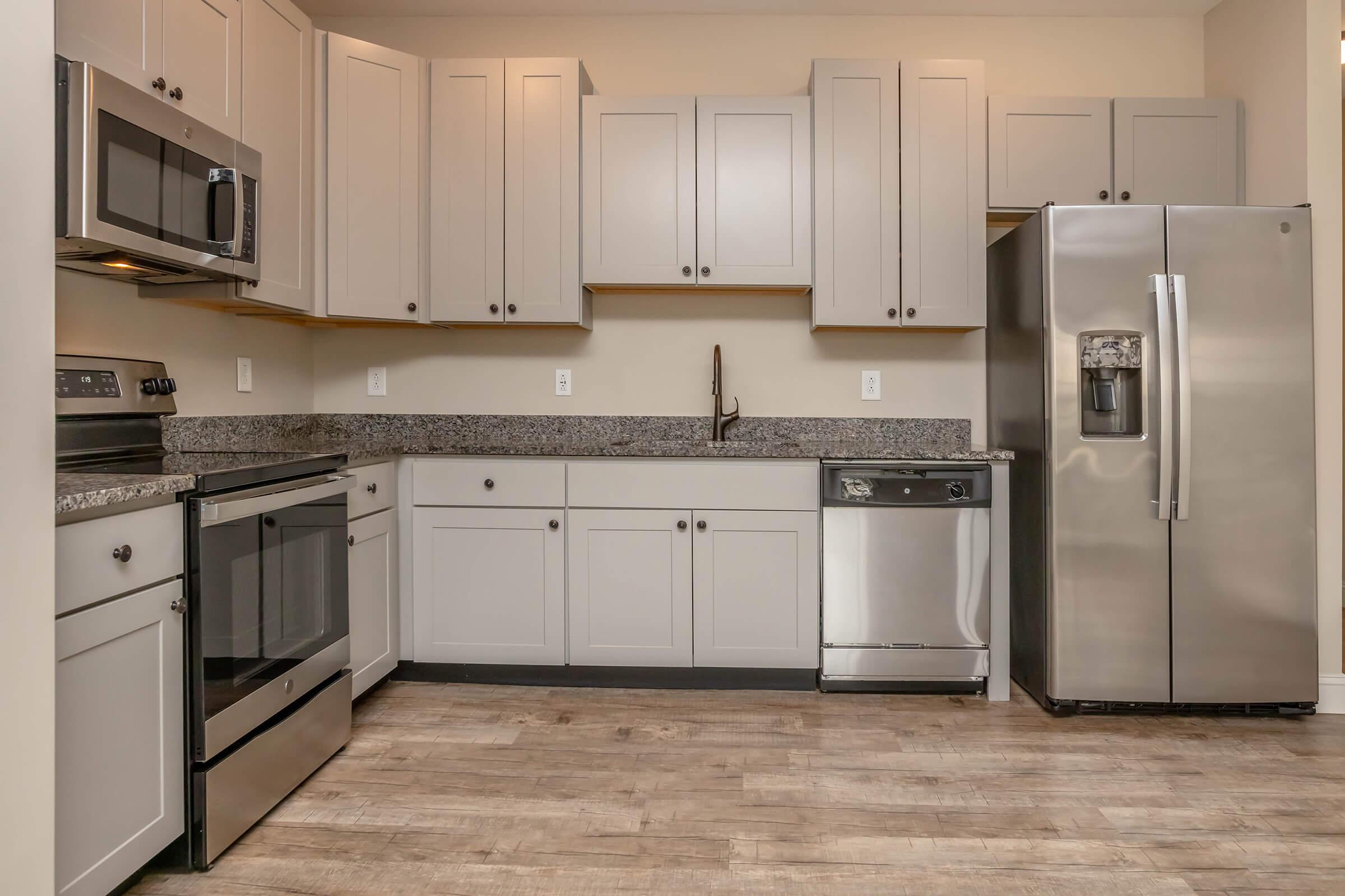 Modern kitchen featuring light gray cabinetry, stainless steel appliances including a microwave, oven, refrigerator, and dishwasher, along with a granite countertop and a wooden floor. The design is clean and contemporary, creating an inviting cooking space.