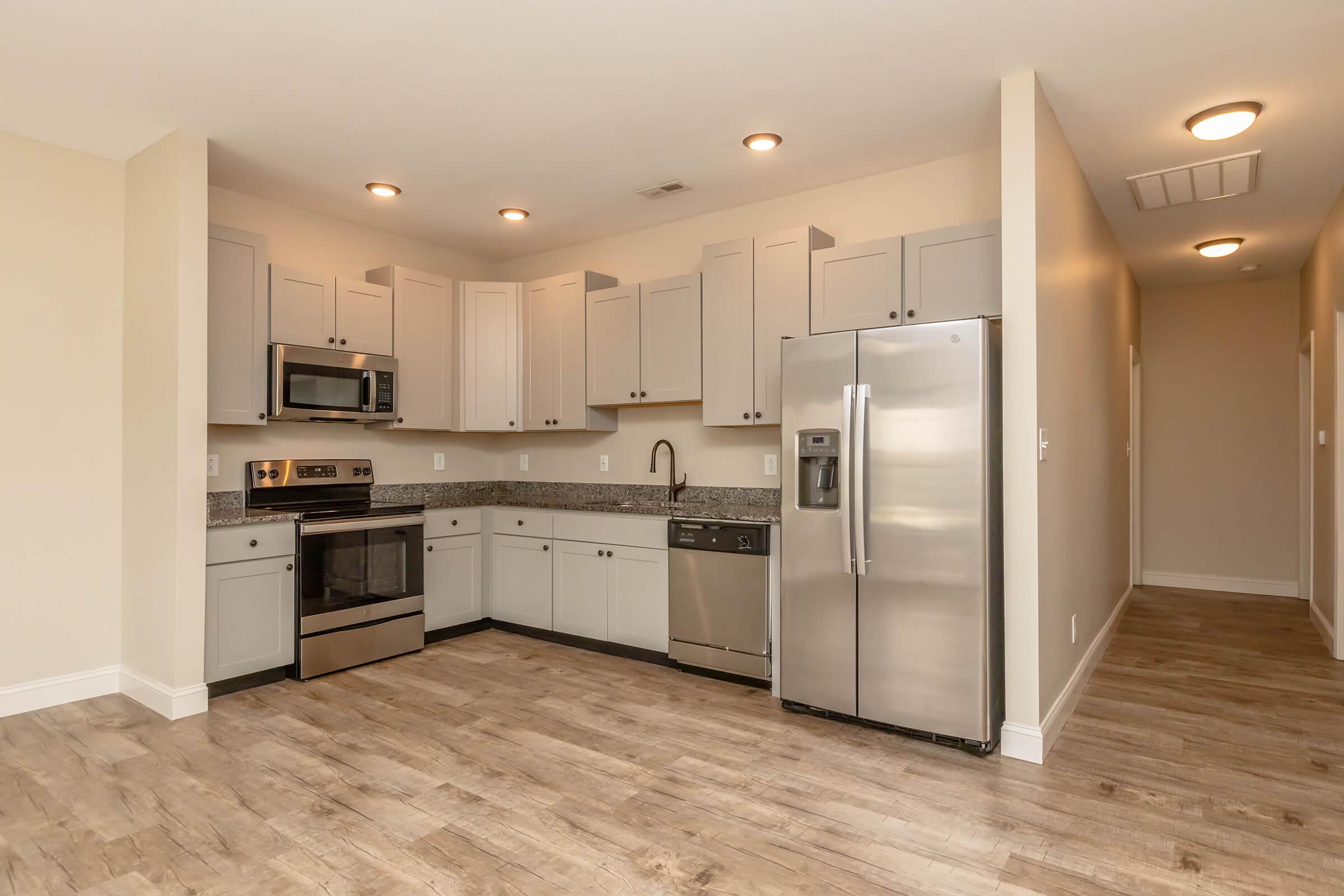 Modern kitchen in an apartment featuring stainless steel appliances, including a microwave, oven, refrigerator, and dishwasher. The cabinetry is light-colored with a granite countertop. The floor is a warm wood-like laminate, and the space is well-lit with recessed lighting.