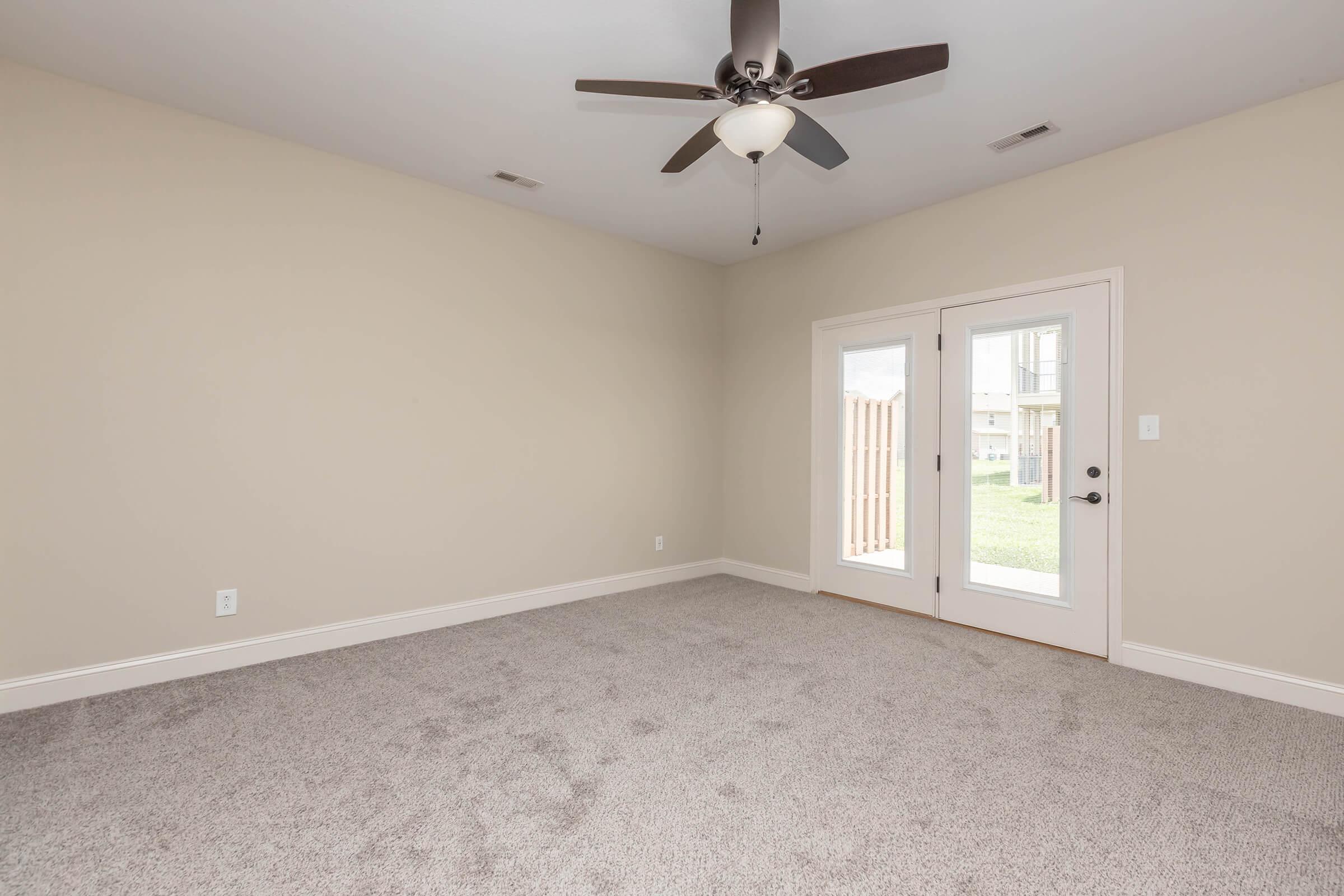 A spacious, empty room with light beige walls and carpeted flooring. It features a ceiling fan, two large windows, and a set of French doors leading outside, allowing natural light to enter. The overall atmosphere is bright and inviting, perfect for personal customization.