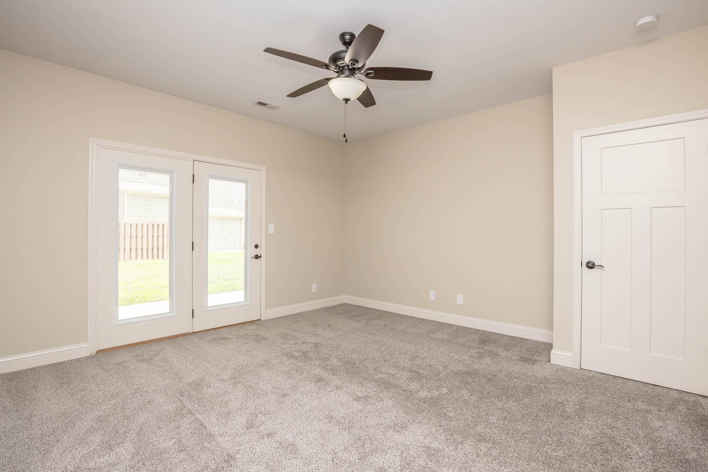 Empty room with light beige walls and plush gray carpet. Features a ceiling fan with dark blades, a pair of glass double doors leading to an outdoor area, and a single door to the right. Natural light enters through the doors, creating a bright and spacious atmosphere.